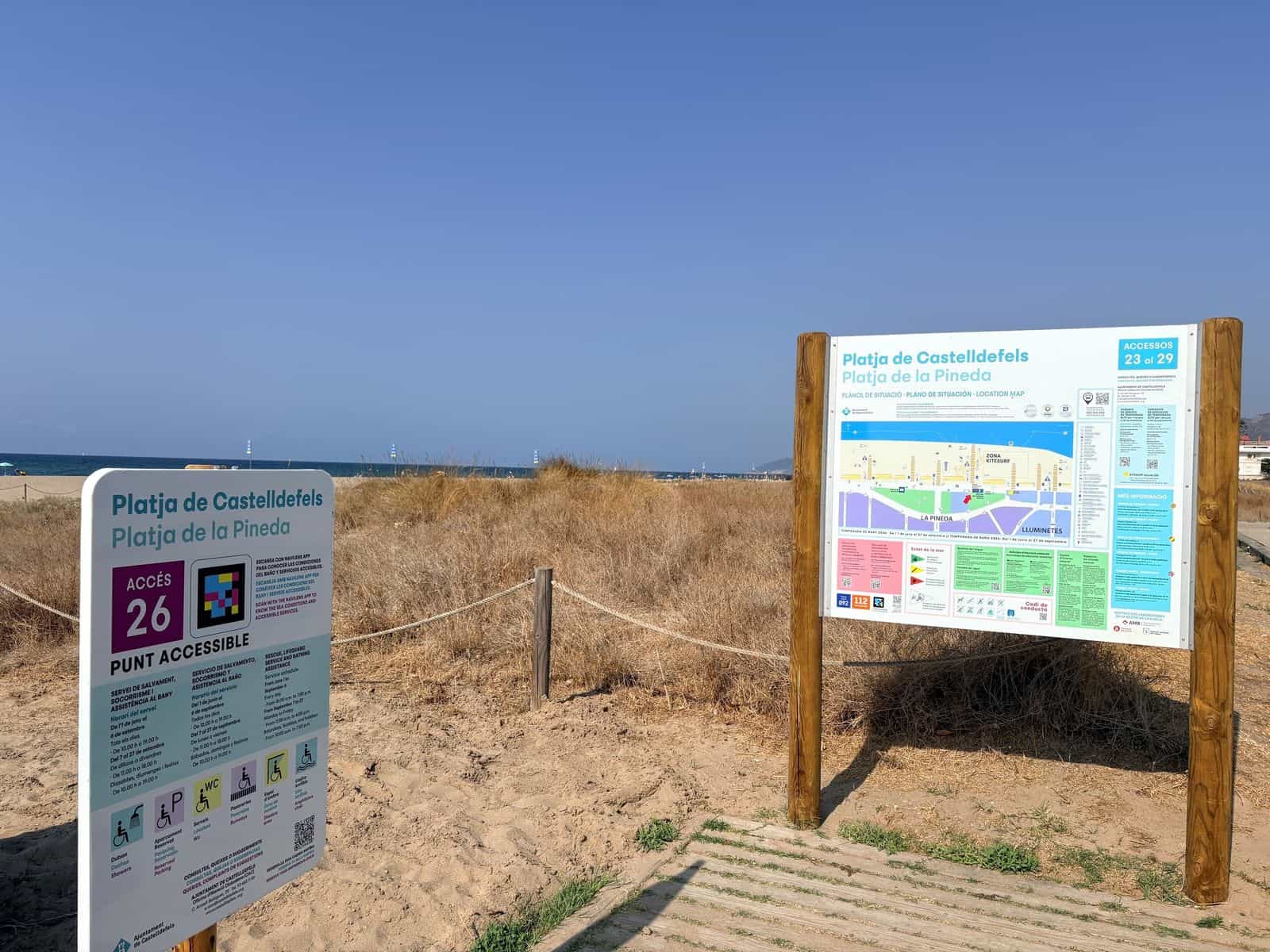 Zugang 26 der Platja de la Pineda in Castelldefels: links das weiße Schild „PUNT ACCESSIBLE“ mit der Nummer 26 und seinem NaviLens-Code; rechts, auf Holzpfosten, die große Tafel „Platja de Castelldefels · Platja de la Pineda — ACCESSOS 23 al 29“ mit dem Lageplan, dahinter Dünen, Sand und Meer