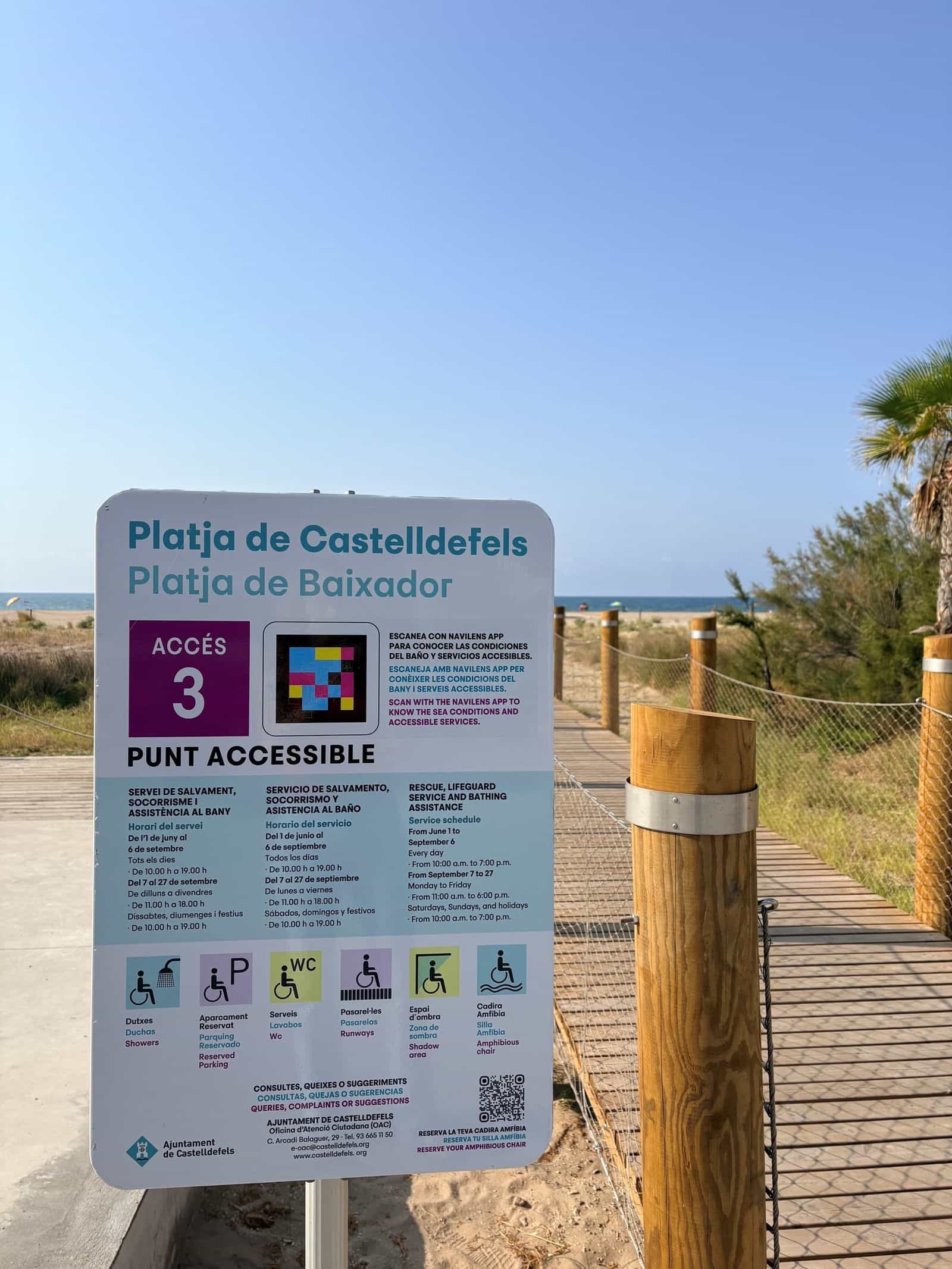 Schild am Zugang 3 der Platja de Baixador in Castelldefels: die violette Marke „ACCÉS 3“, der NaviLens-Code in Cyan, Magenta und Gelb und daneben der dreisprachige Text „SCAN WITH THE NAVILENS APP TO KNOW THE SEA CONDITIONS AND ACCESSIBLE SERVICES“, mit den Zeiten des Rettungsdienstes, den sechs Service-Piktogrammen und dahinter dem Holzsteg, der die Dünen zum Ufer hin durchquert