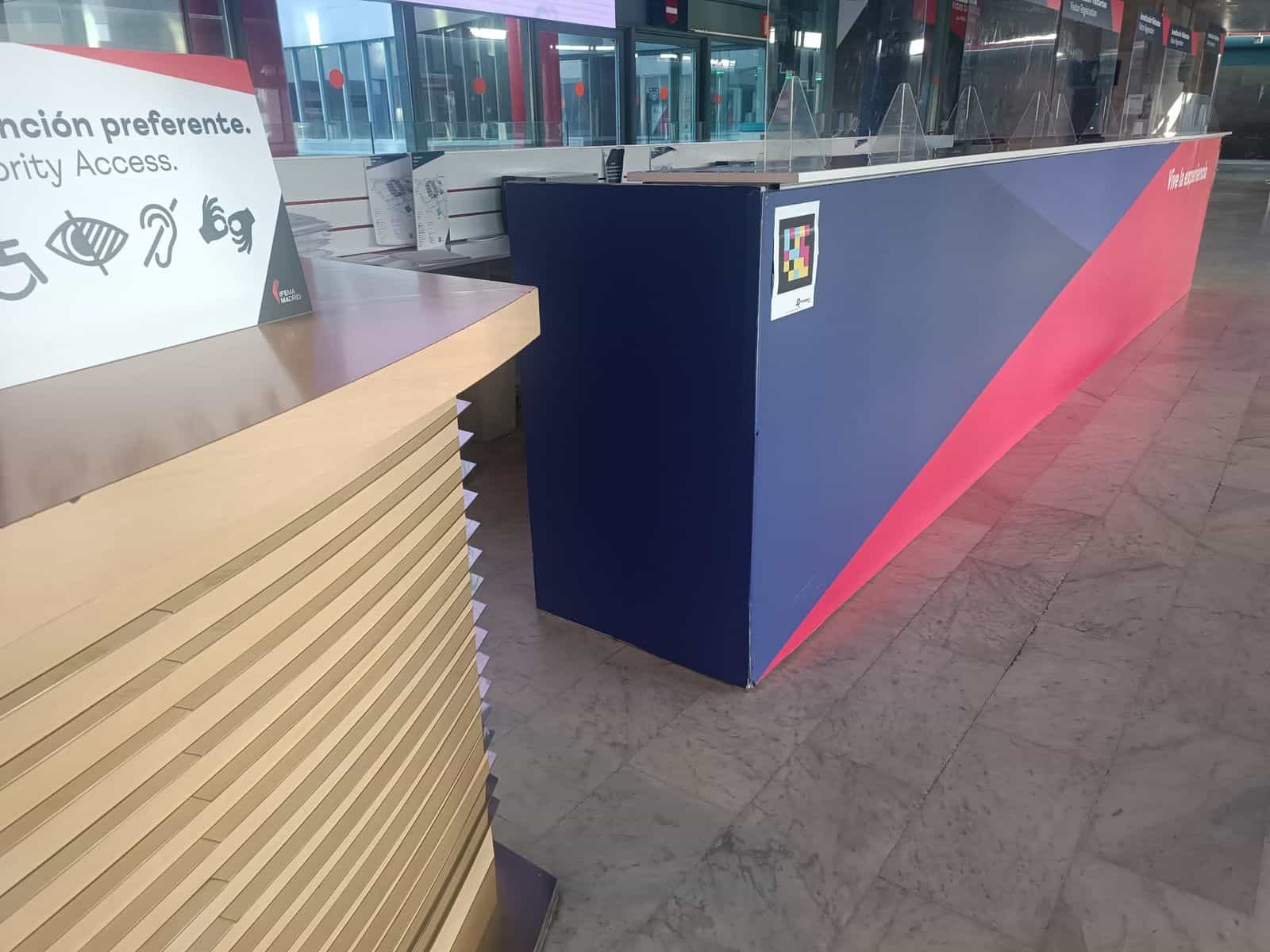 Banco degli accrediti di IFEMA MADRID con grafica azzurra e rossa: a sinistra il cartello «Accesso prioritario / Priority Access» con pittogrammi di disabilità; a destra un codice NaviLens quadrato applicato sul fianco del banco