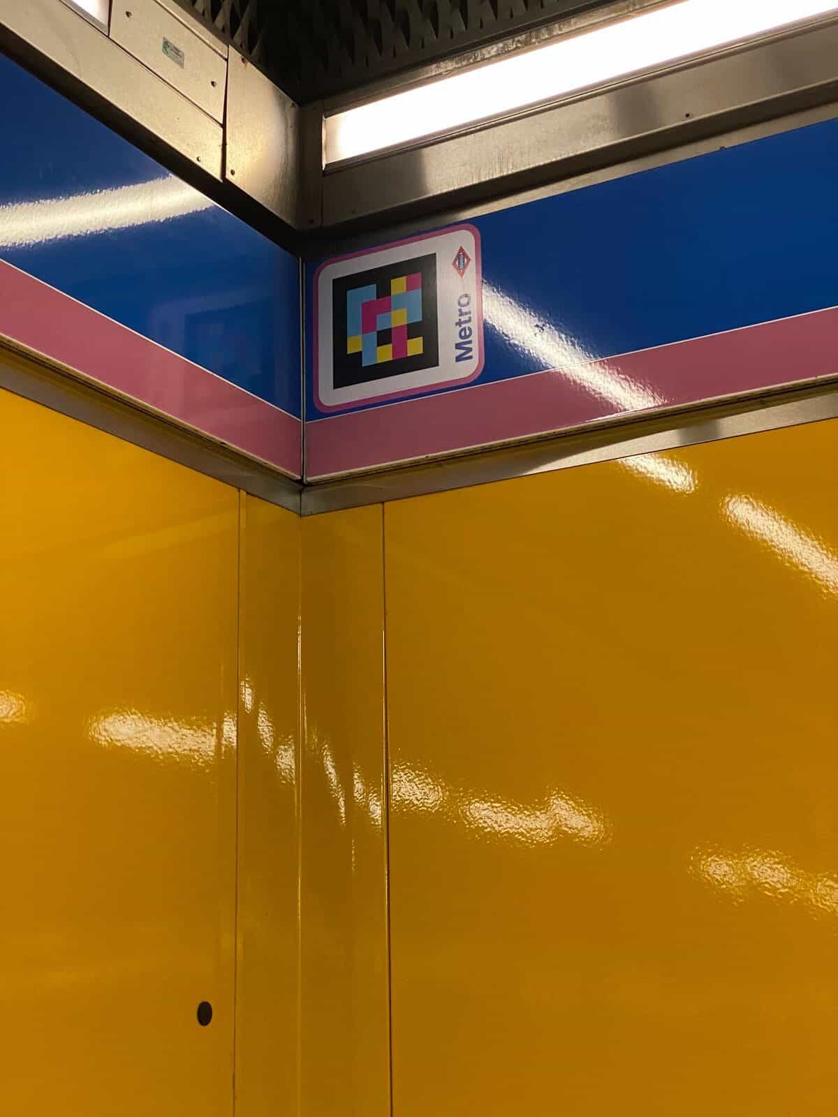 Detail einer leuchtend gelben Ecke des Bahnhofs Flughafen T1-T2-T3: oben an der blau-rosa Kante der Linie 8 der gerahmte NaviLens-Code neben der roten Metro-Raute