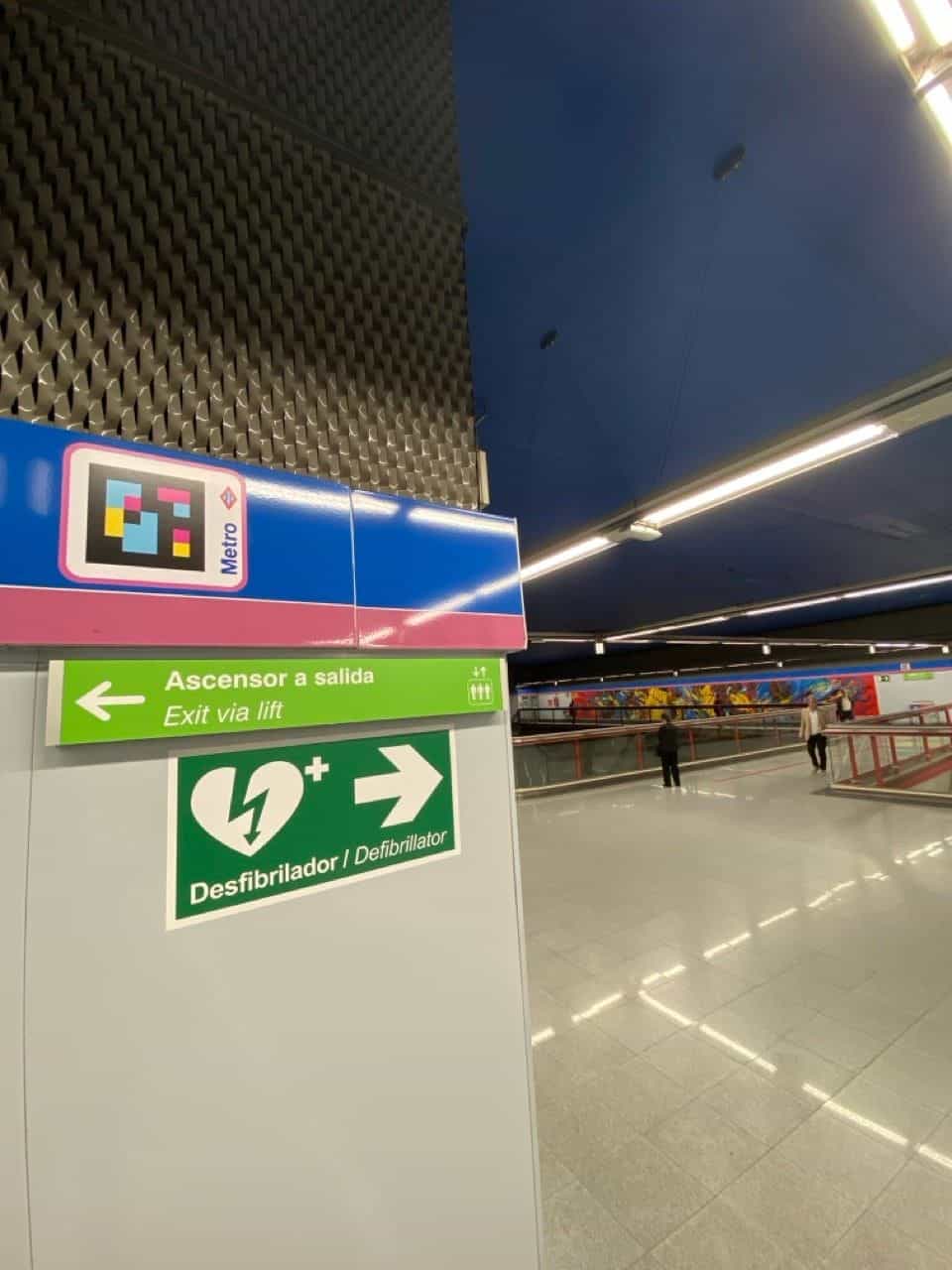 Verteilerebene des Bahnhofs Flughafen T1-T2-T3: an der Oberkante über dem blau-rosa Band der Linie 8 ein NaviLens-Code neben der roten Metro-Raute; darunter das grüne Schild „Aufzug zum Ausgang“ und „Defibrillator“; im Hintergrund der Korridor mit einem farbenfrohen künstlerischen Wandbild, das den Bahnhof schmückt