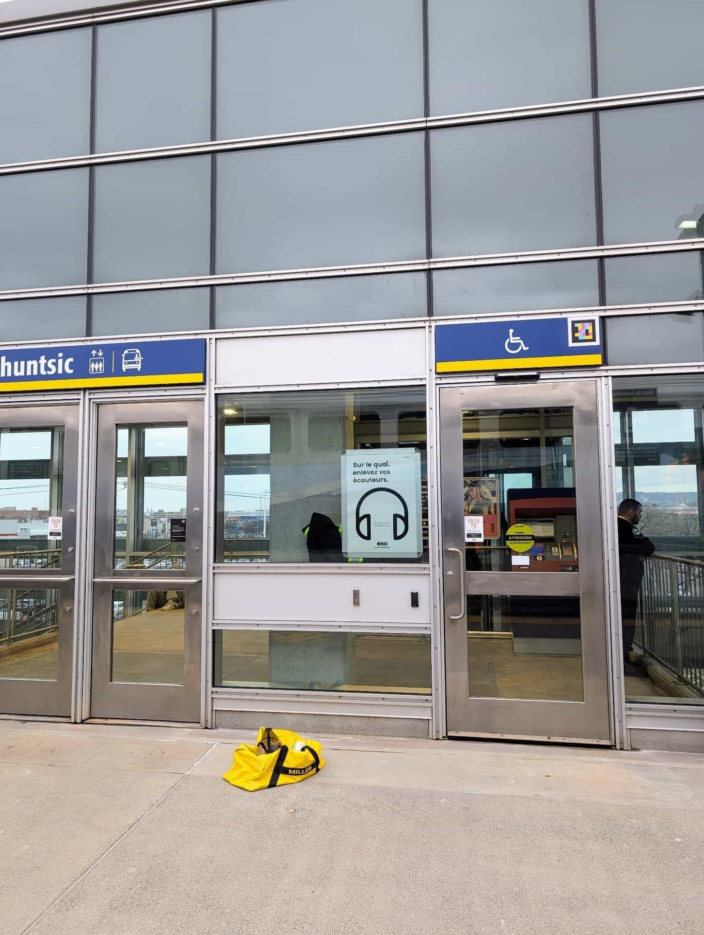 Twin access to Gare Ahuntsic on a glazed elevated walkway: blue-and-yellow “Ahuntsic” sign with elevator and bus pictograms and, on the right-hand door marked with the wheelchair pictogram, the NaviLens code