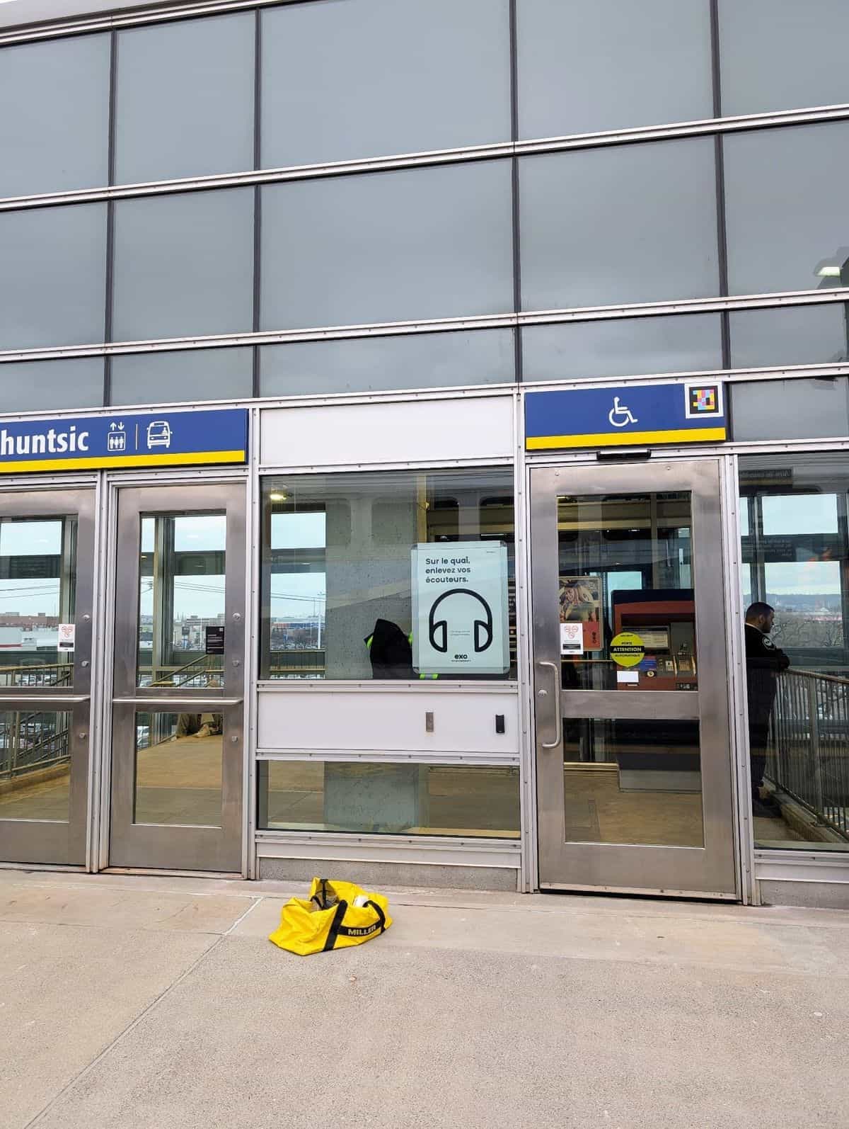 Twin access to Gare Ahuntsic on a glazed elevated walkway: blue-and-yellow «Ahuntsic» sign with elevator and bus pictograms and, on the right-hand door marked with the wheelchair pictogram, the NaviLens code