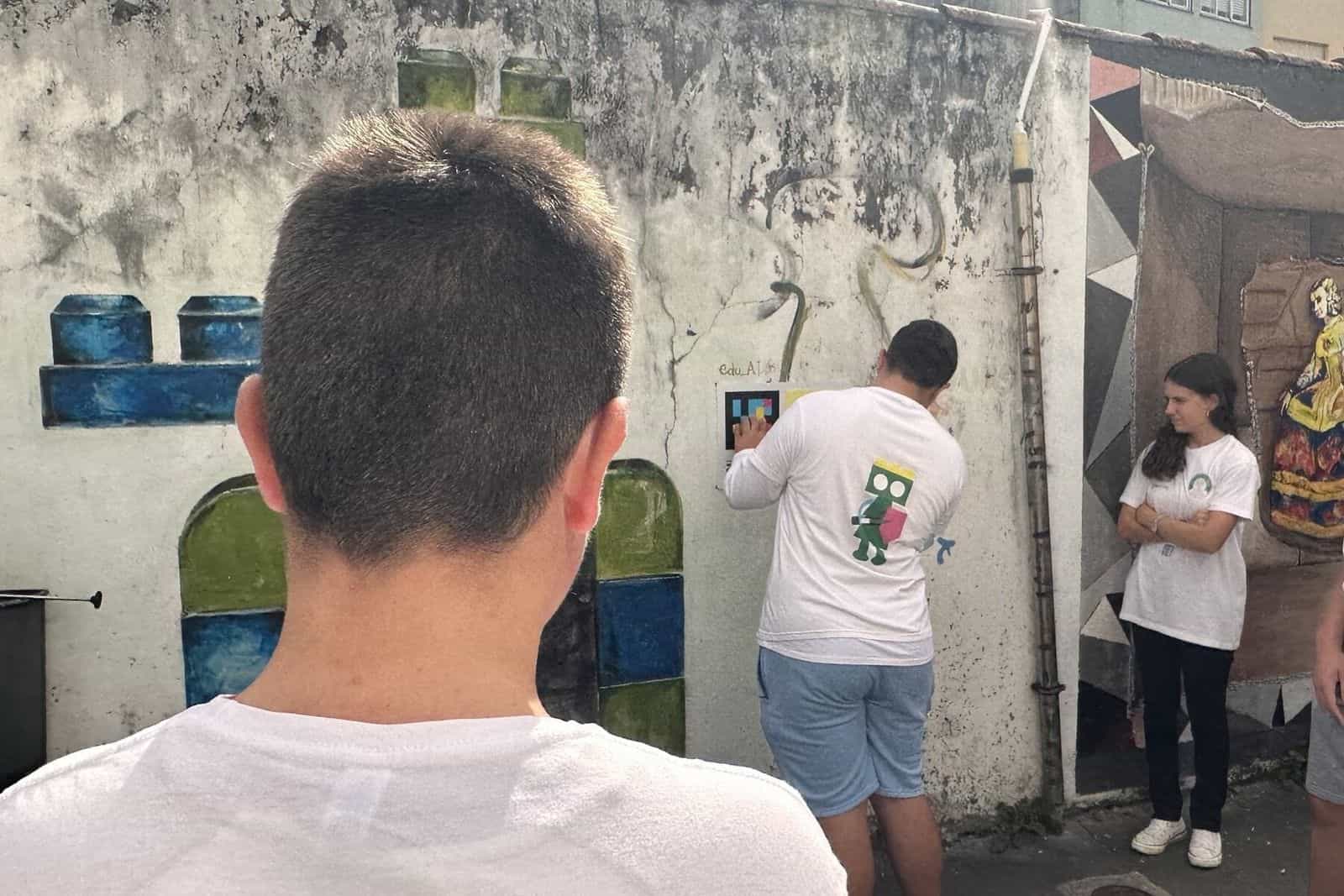 Studente dell’IES di Fene che scansiona col telefono un pannello «Tocando as Meninas» fissato a un muro di Canido accanto a un murale dipinto in blu, verde e bianco