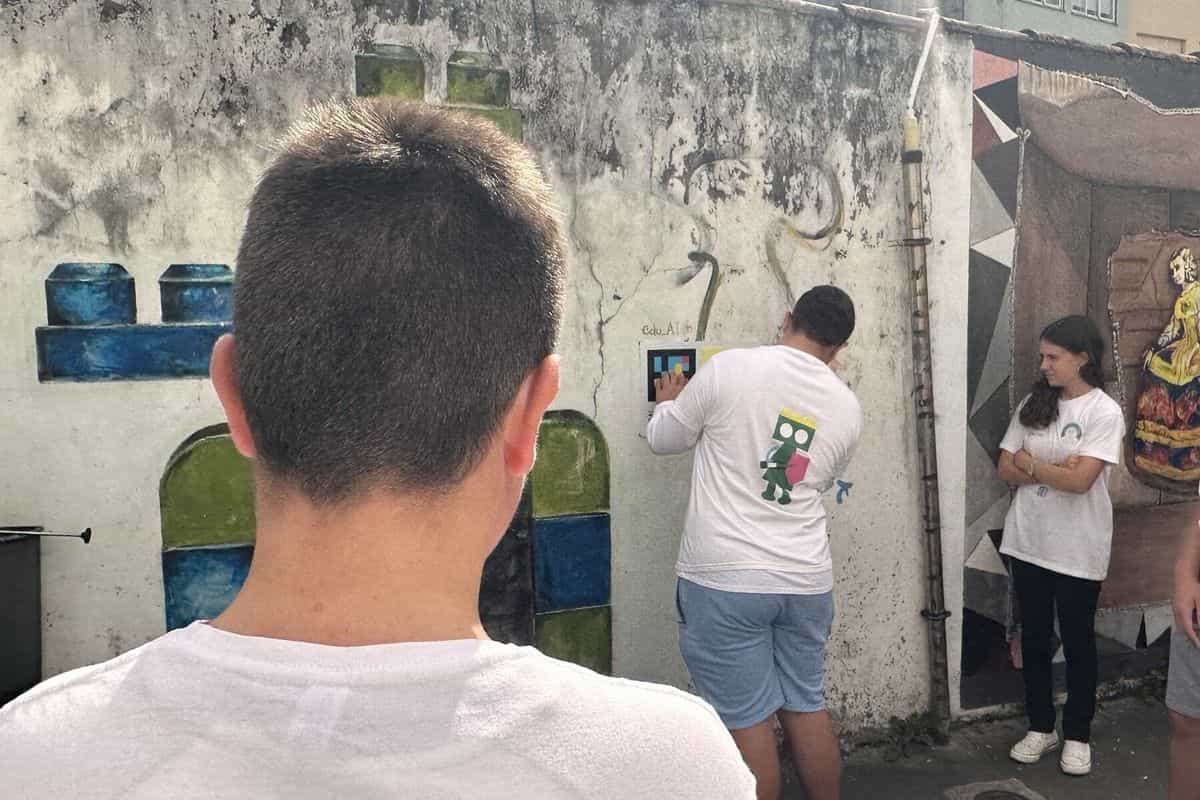 Alumno del IES de Fene escaneando con su móvil un panel «Tocando as Meninas» fijado a un muro de Canido junto a un mural pintado en azul, verde y blanco