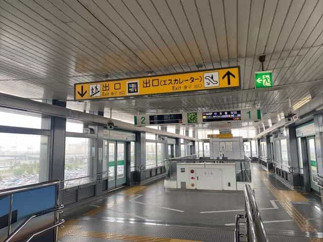 神戸空港駅の中間コンコース。黄色い吊り下げサイン「出口 Exit」と「出口(エスカレーター)」にそれぞれNaviLensタグ。奥には1番線・2番線の緑色ホームドアと黄色の点字ブロック