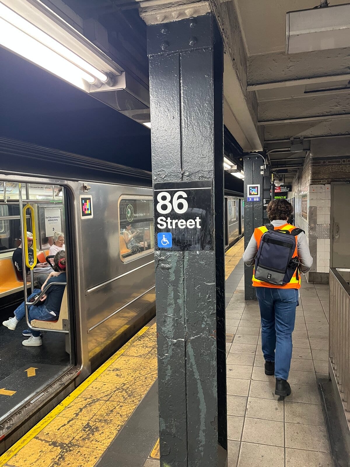 Quai de 86 Street (lignes 4-5-6, Lexington Av) : colonne peinte en noir avec le panneau blanc «86 Street» et l'icône bleue d'accessibilité ; en haut, un marqueur coloré NaviLens ; au premier plan, un membre de l'équipe NaviLens en gilet orange réfléchissant marchant sur le quai