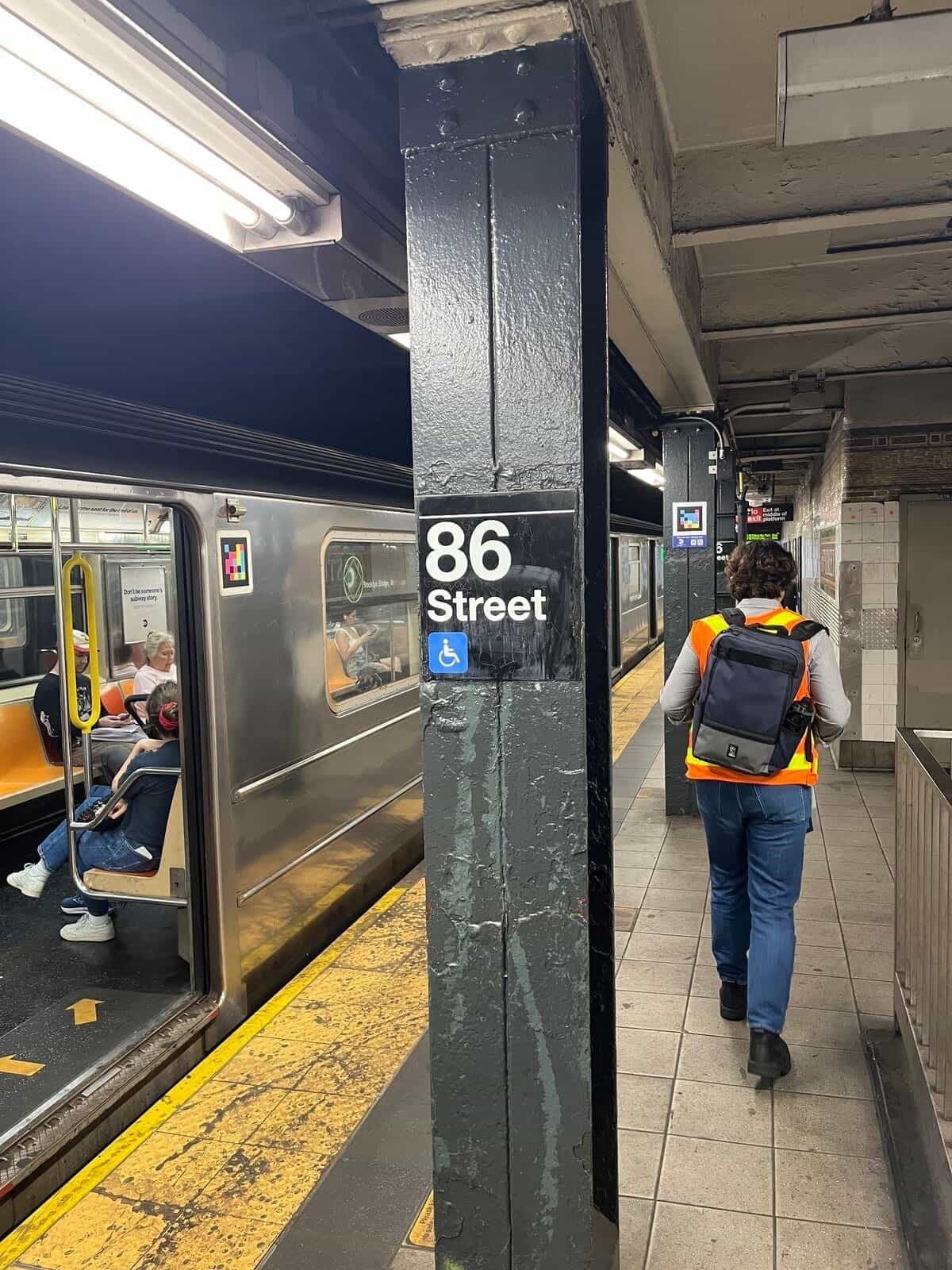 Andén de 86 Street (líneas 4-5-6, Lexington Av): columna pintada en negro con el rótulo blanco «86 Street» y el icono azul de accesibilidad; arriba, un código NaviLens de cuadros de colores; en primer plano, una persona del equipo NaviLens con chaleco naranja reflectante caminando por el andén