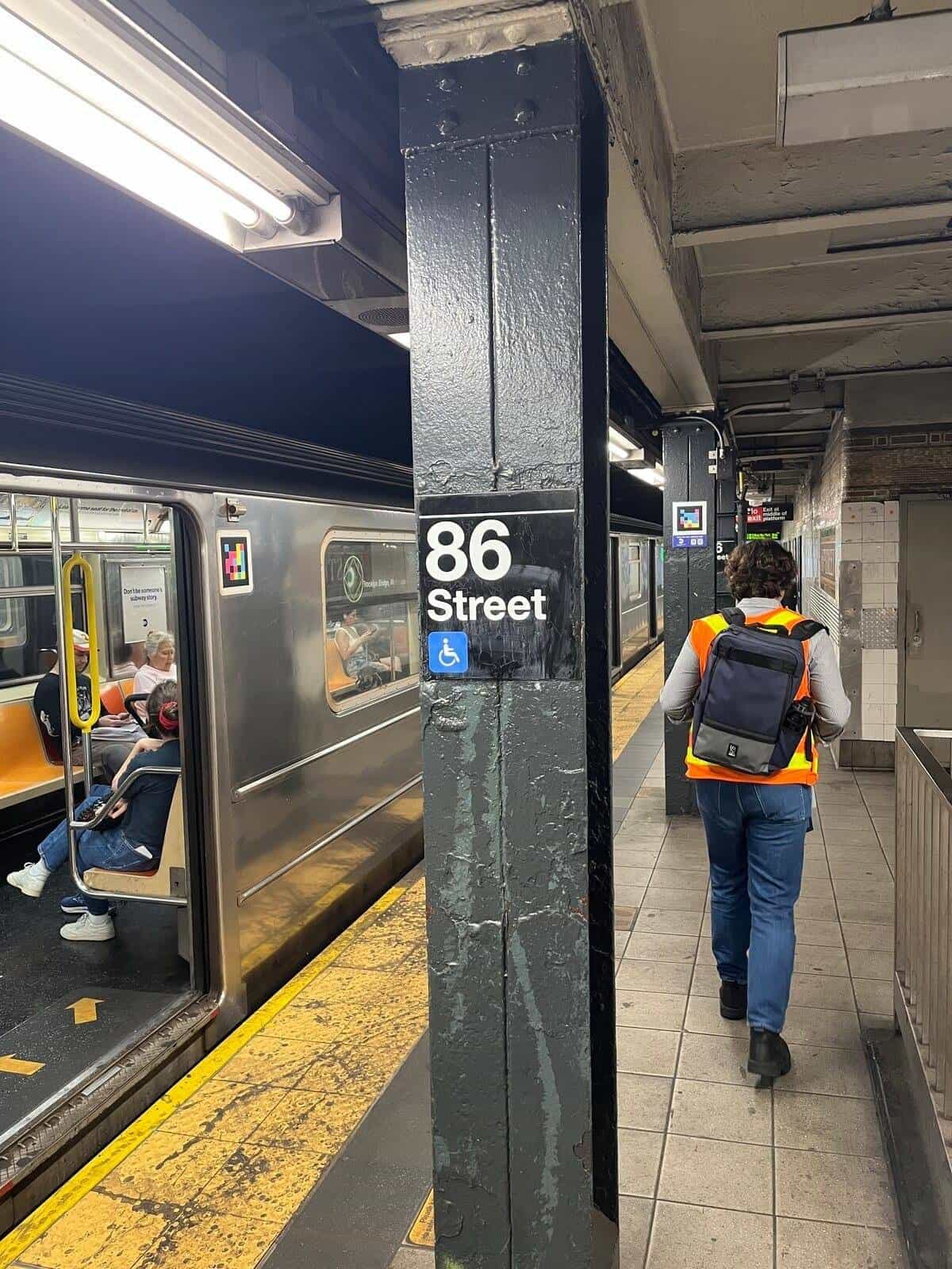 86 Street platform (4-5-6, Lexington Av): black-painted column with the white “86 Street” roundel and the blue accessibility icon; up top, a NaviLens colour-coded marker; in the foreground, a NaviLens team member in a high-vis orange vest walking along the platform