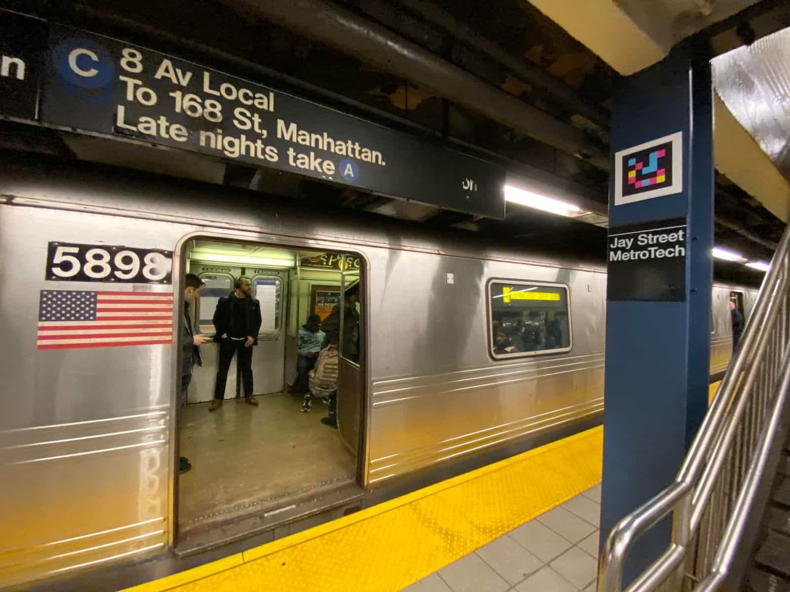 C-line train stopped at Jay St-MetroTech: silver car no. 5898 with US flag and the sign «C · 8 Av Local · To 168 St, Manhattan. Late nights take A». To the right, a blue column with the «Jay Street MetroTech» sign and a square NaviLens code on top