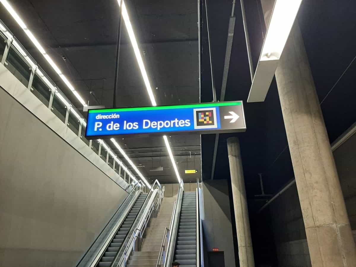 Cartel luminoso azul «dirección P. de los Deportes» con código NaviLens y flecha sobre las escaleras mecánicas