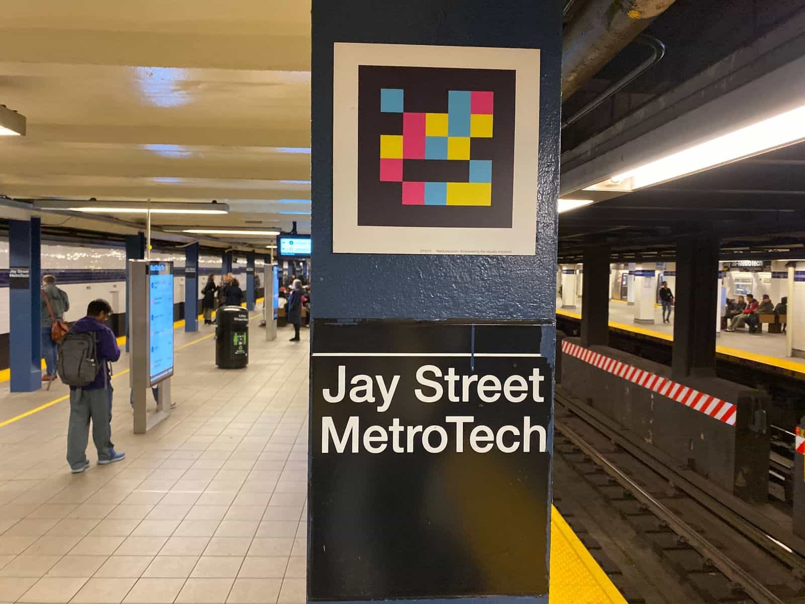 Platform at Jay St-MetroTech: dark blue painted column with a square NaviLens code and, below, the black-on-white “Jay Street MetroTech” sign. In the background, riders checking the digital subway information panels