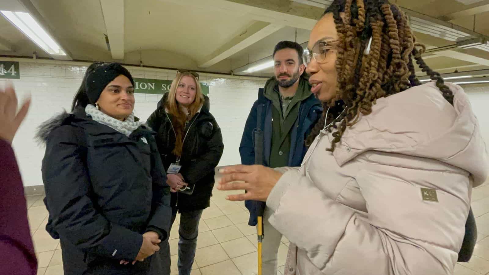 Low-vision user gestures while explaining something to the team on the line-4 platform at Union Square; in the background, the green sign with the “4” numeral and the white-tile wall typical of the station