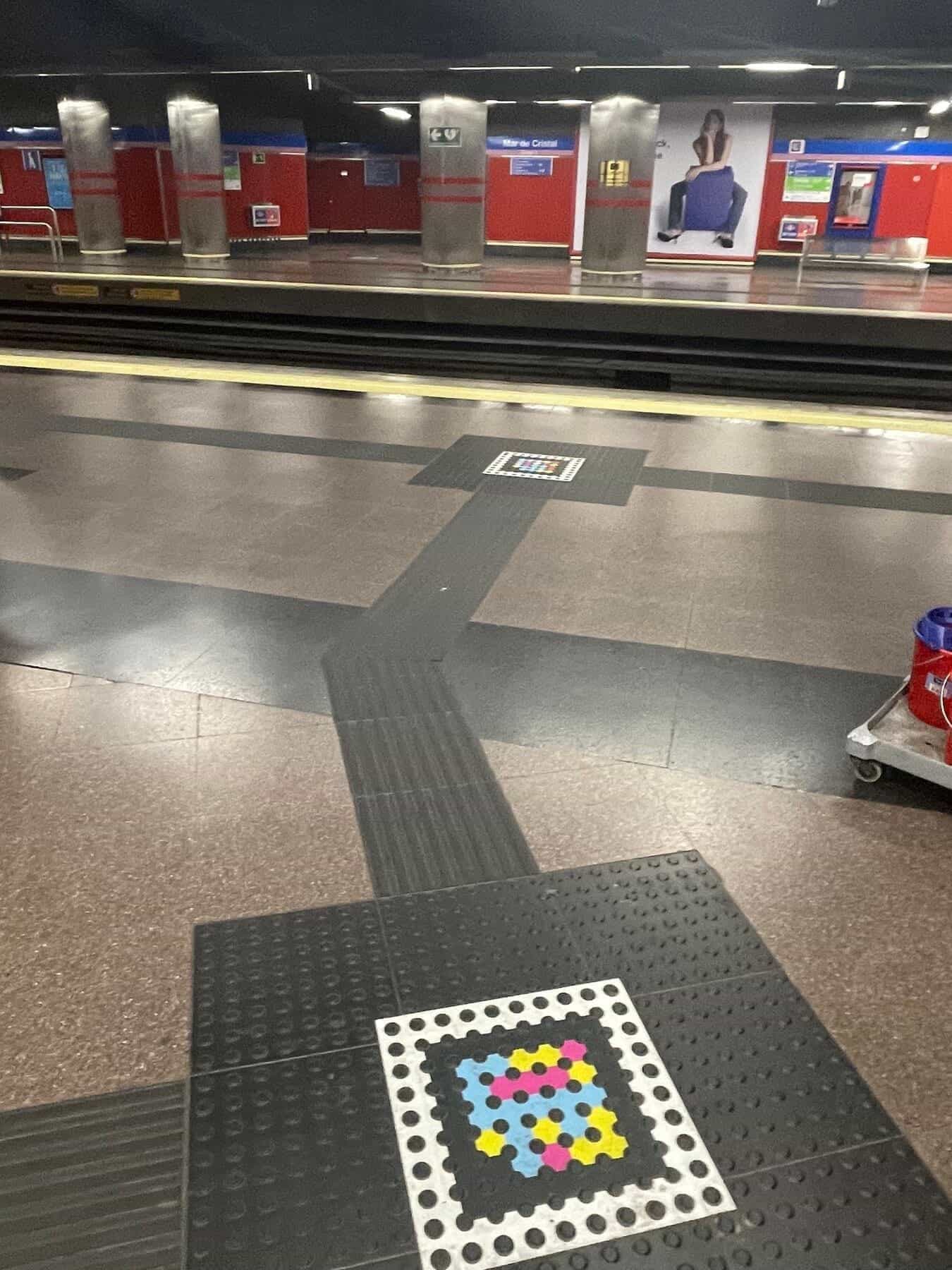 Bodendetail des Bahnsteigs Mar de Cristal: ein Quadrat aus Keramikfliesen – rot, gelb, blau, weiß und schwarz – gibt das NaviLens-Code-Muster wieder und fügt sich in den grauen, gerillten Blindenleitbelag ein; im Hintergrund ein weiß-blauer Metro-Madrid-Zug am gegenüberliegenden Bahnsteig