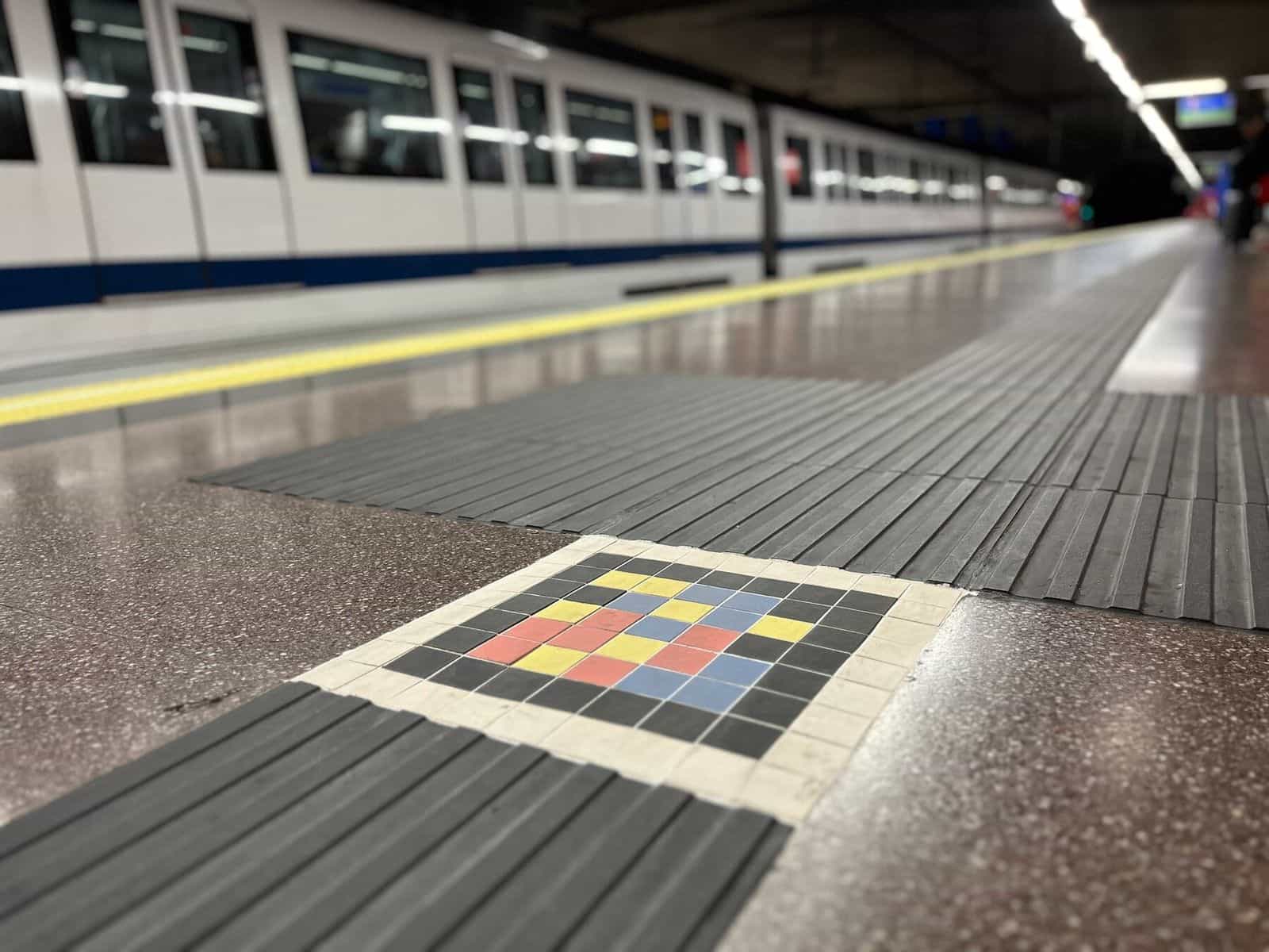 Bahnsteig Mar de Cristal mit einem weiß-blauen Metro-Madrid-Zug im Hintergrund; im Vordergrund, eingelassen in den grauen gerillten Blindenleitbelag, ein Quadrat aus gelben, roten, blauen und schwarzen Keramikfliesen, das den NaviLens-Code wiedergibt
