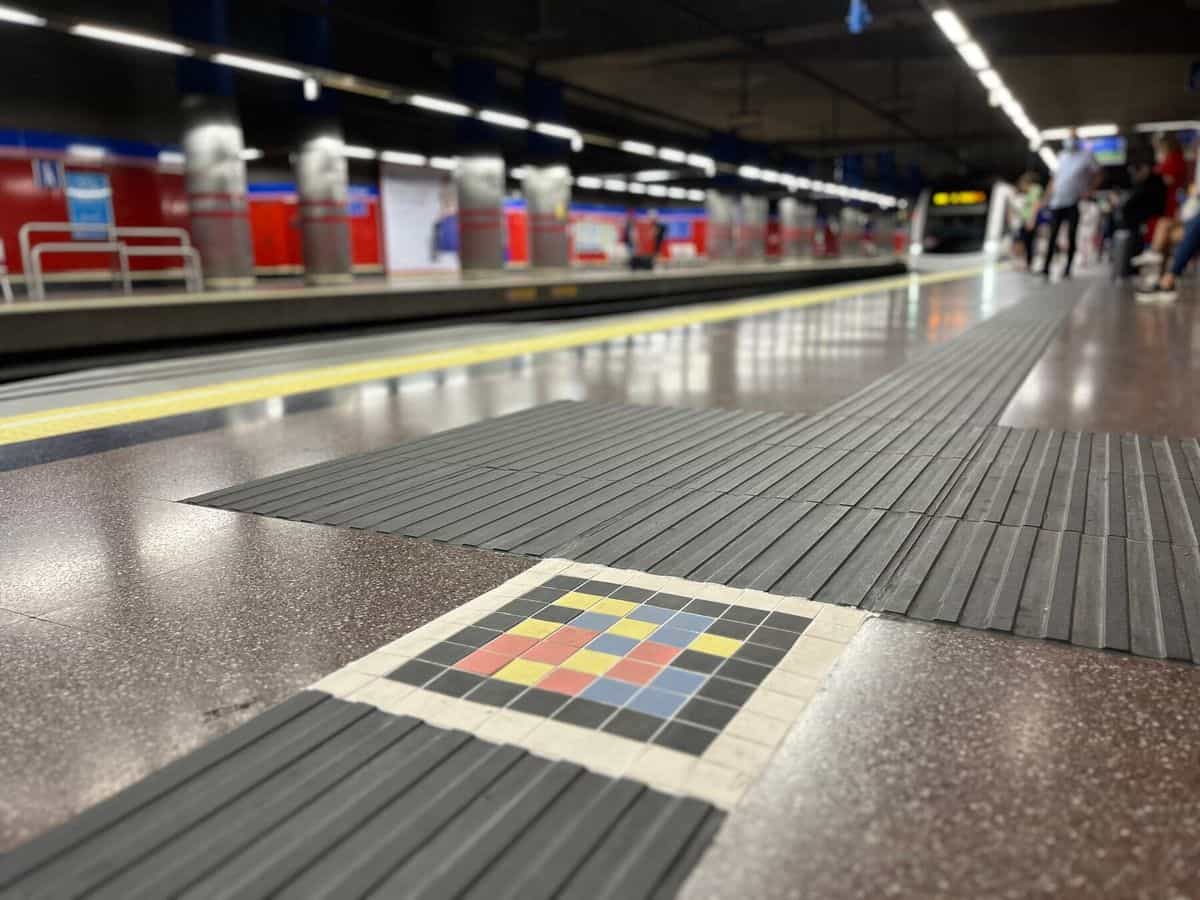 Bodenansicht des Bahnsteigs Mar de Cristal: der graue Blindenleitbelag durchquert den glänzenden Granitboden bis zur gelben Sicherheitslinie, an seiner Kreuzung ein quadratisches Mosaik aus farbigen Keramik-Tesserae mit dem NaviLens-Code; im Hintergrund warten Fahrgäste an den Stahlpfeilern und den charakteristischen roten Wänden