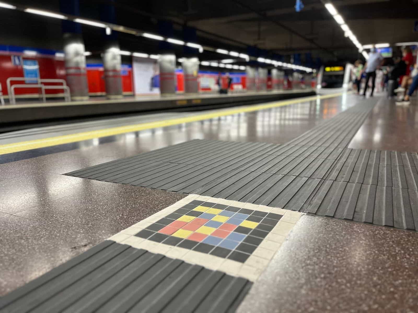 Bodenansicht des Bahnsteigs Mar de Cristal: der graue Blindenleitbelag durchquert den glänzenden Granitboden bis zur gelben Sicherheitslinie, an seiner Kreuzung ein quadratisches Mosaik aus farbigen Keramik-Tesserae mit dem NaviLens-Code; im Hintergrund warten Fahrgäste an den Stahlpfeilern und den charakteristischen roten Wänden