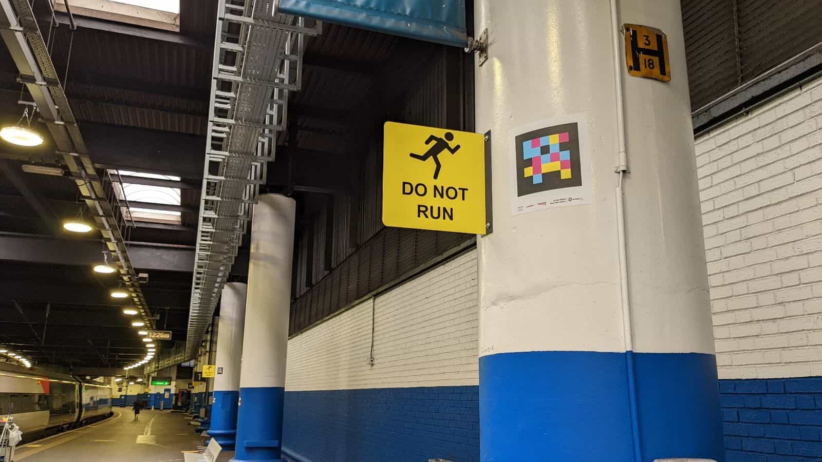Euston platform with white-and-blue pillar, yellow «DO NOT RUN» sign, pillar marker «3/18 H» and large NaviLens code with «SCAN FROM 14M FAR AWAY» pictogram