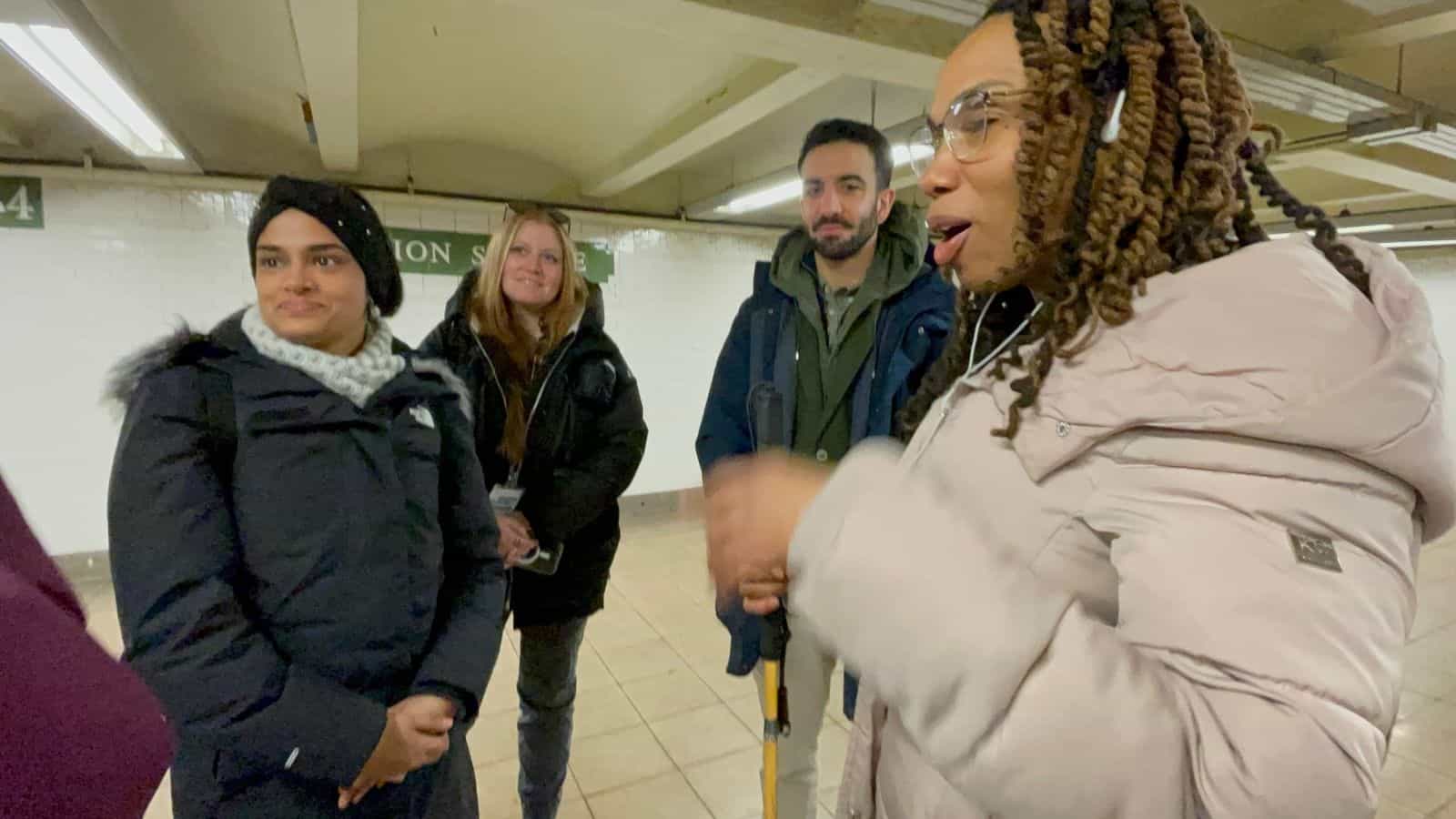 Group listening to a white-cane user on the platform at Union Square; in the background, the green sign reading “Union Sq” in white capitals