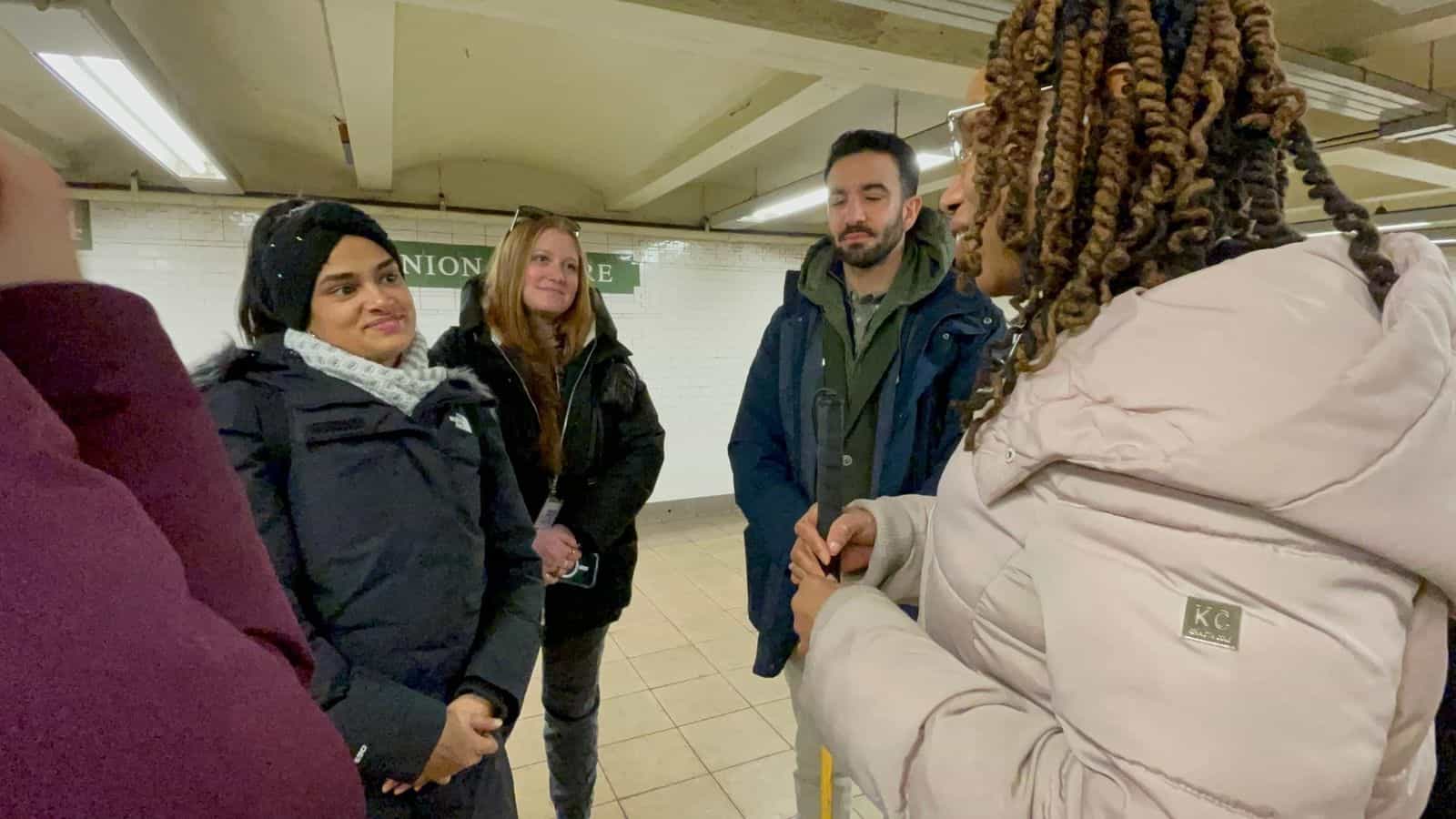 Team conversation on the platform at Union Square: on the left, the green station sign; in the background, the white tiles typical of the NYC subway