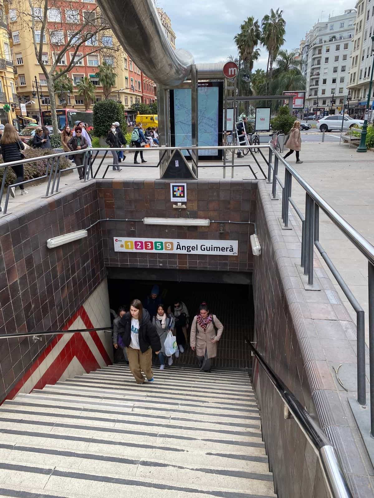 Boca de acceso a la estación Àngel Guimerà con el rótulo '1 2 3 5 9 Àngel Guimerà' a colores sobre cerámica marrón; sobre el dintel, un código NaviLens cuadrado a colores; varias personas suben las escaleras desde el vestíbulo