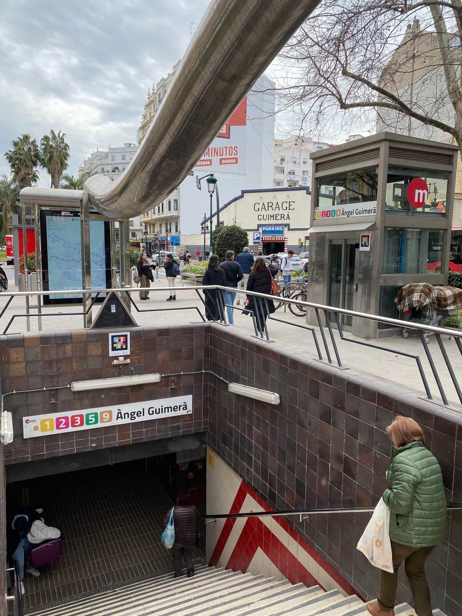 Veduta verso la strada dalla cima della scala d’accesso ad Àngel Guimerà, con la facciata del «Garage Guimerà» e un parcheggio in fondo; parapetti cromati, pedoni che attraversano e biciclette parcheggiate a destra