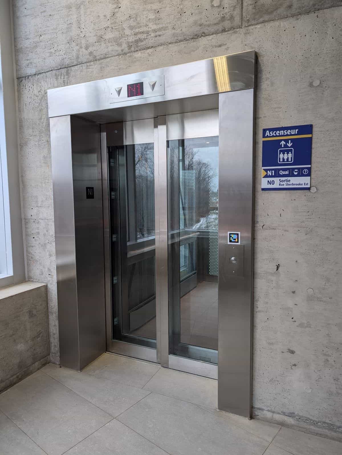 Panoramic stainless-steel and glass elevator at an exo station: blue-and-yellow «Ascenseur» sign reading «N1 Quai · N0 Sortie Rue Sherbrooke Est»; below the call button, a square NaviLens code in cyan, magenta and yellow