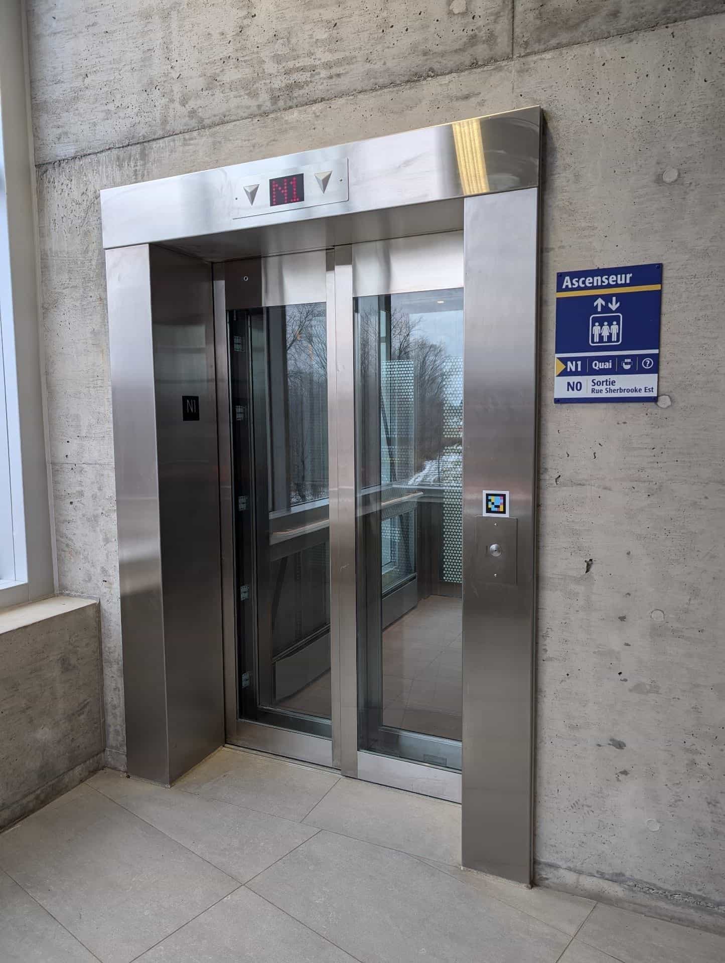 Panoramic stainless-steel and glass elevator at an exo station: blue-and-yellow “Ascenseur” sign reading “N1 Quai · N0 Sortie Rue Sherbrooke Est”; below the call button, a square NaviLens code in cyan, magenta and yellow