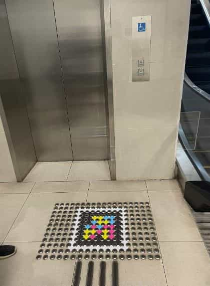 Ascensor accesible del interchange: puerta metálica gris, pictograma azul de silla de ruedas en la pared, botonera braille y, en el suelo frente a la puerta, código NaviLens enmarcado por una alfombra de botones podotáctiles; a la derecha se intuye una escalera mecánica