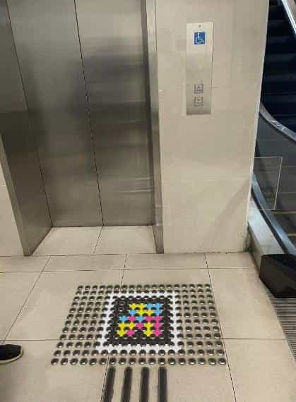 Accessible lift inside the interchange: grey metal door, blue wheelchair pictogram on the wall, braille buttons and, on the floor in front of the door, a NaviLens code framed by a mat of tactile dots; on the right, an escalator can be glimpsed