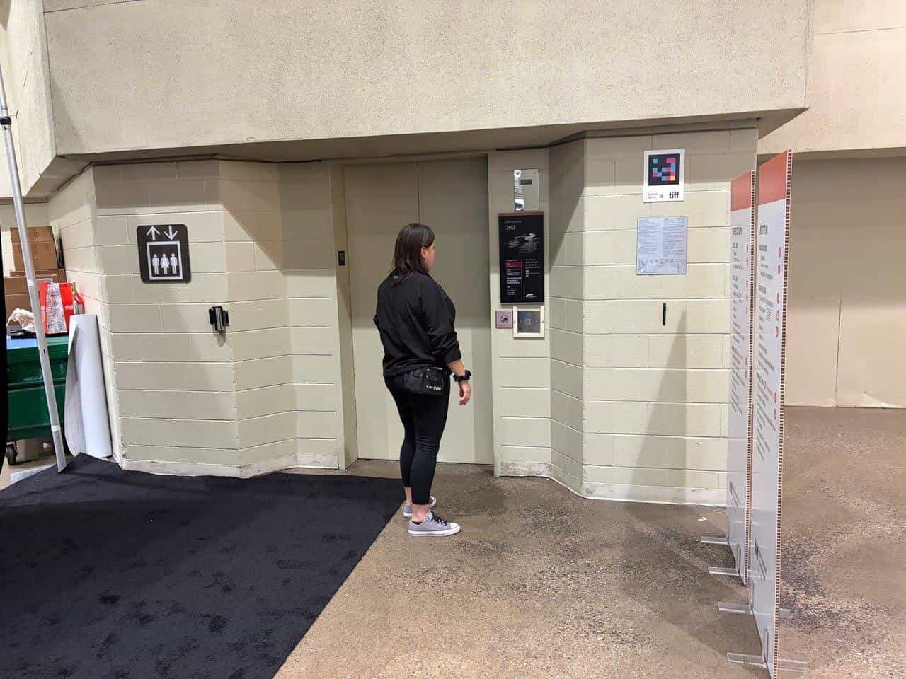 A delegate waits at the level 300 lift in the Metro Toronto Convention Centre. To the right of the door, on the pale block wall, a NaviLens code with the TIFF logo, above the printed venue map.