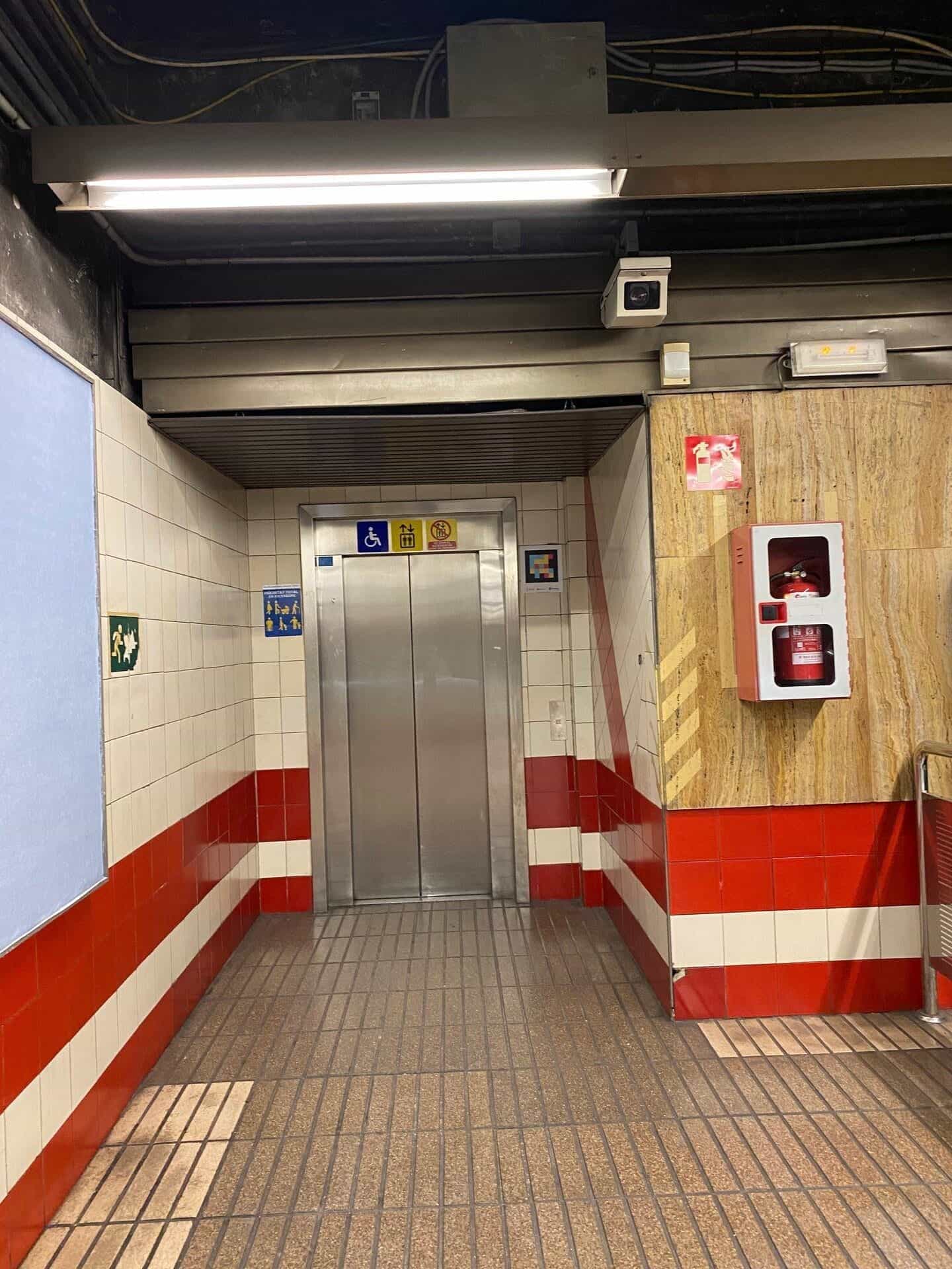 Vano ascensore della banchina con porta metallica, pittogrammi di priorità per persone con mobilità ridotta · passeggini · valigie e, a destra, un codice NaviLens quadrato a colori con la firma Generalitat Valenciana · Metrovalencia · NaviLens; in fondo, estintore e fasce rosse caratteristiche della stazione