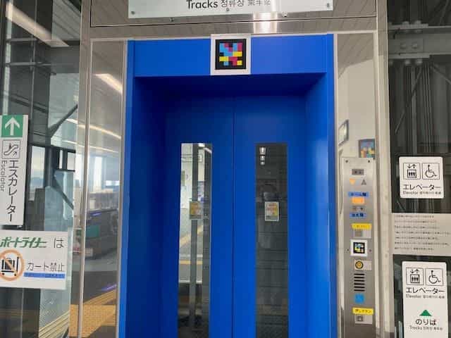神戸空港駅の青いエレベーター扉。鴨居部分に白枠のNaviLensタグ。両側には点字付きの「エレベーター / Elevator」絵文字と「ポートライナーはカート禁止」パネル