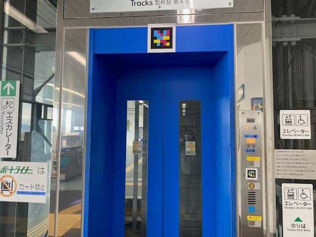 Blue lift door at Kobe Airport Station with a white-framed NaviLens tag on the lintel; on either side, braille “Lift” pictograms and a “No trolleys on the Port Liner” panel