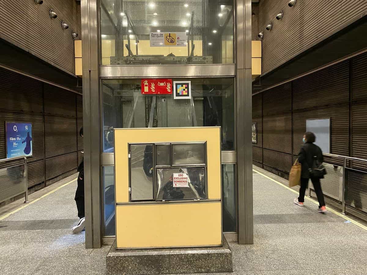 Cabina de ascensor acristalado con armario metálico de uso exclusivo de bomberos en la base, paredes de cristal transparente y, sobre el dintel, un código NaviLens cuadrado a colores junto a los pictogramas de seguridad de la estación