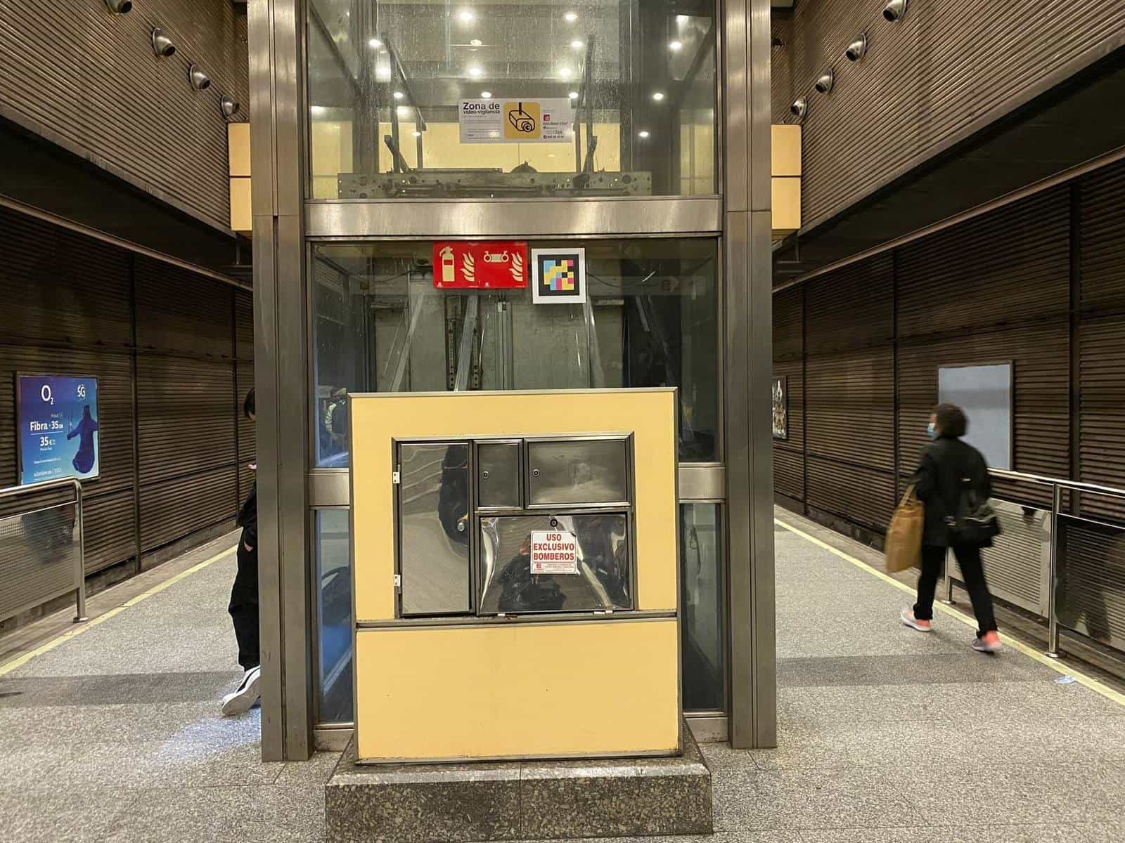 Glass-walled lift cabin with a metal cabinet for firefighters' use only at its base, transparent glass walls and, above the lintel, a colourful square NaviLens code next to the station safety pictograms.