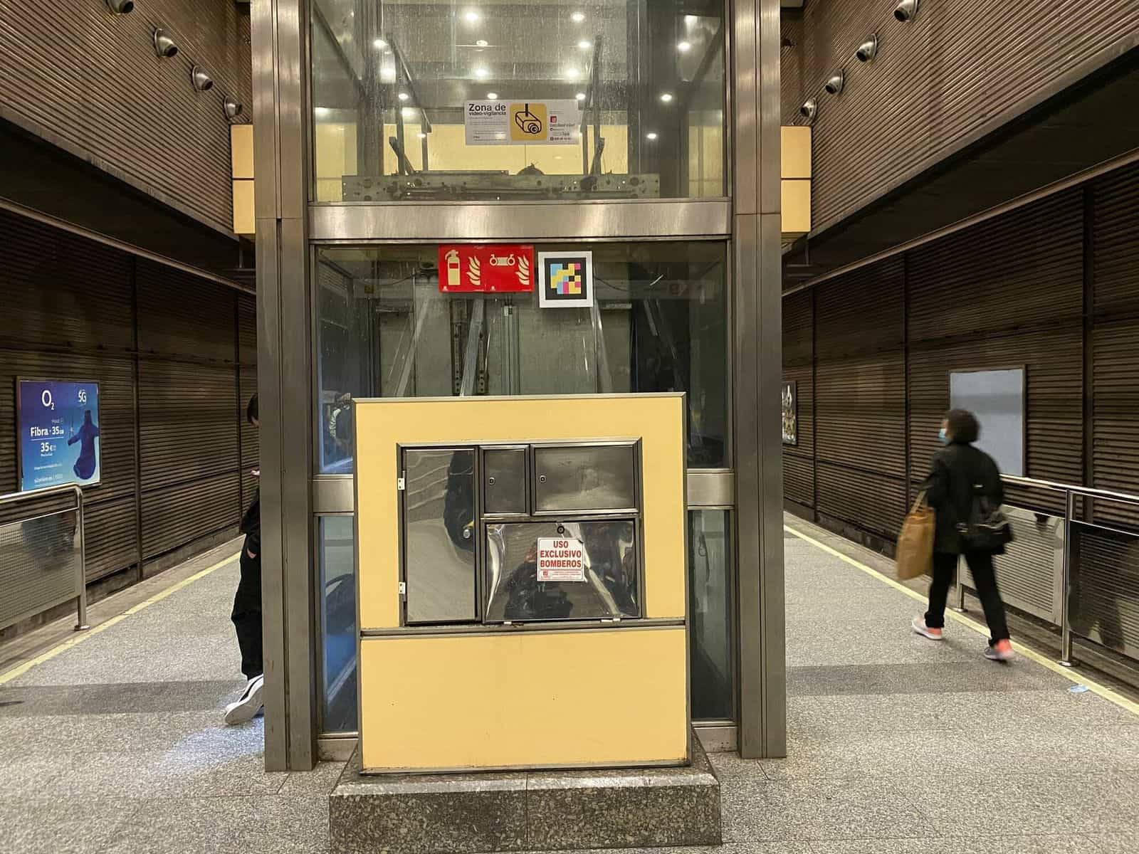 Cabina di ascensore a vetri con armadietto metallico a uso esclusivo dei vigili del fuoco alla base, pareti di vetro trasparente e, sull’architrave, un codice NaviLens quadrato a colori accanto ai pittogrammi di sicurezza della stazione