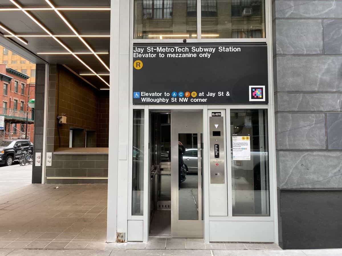 Ascensor exterior de Jay St-MetroTech en la esquina noroeste de Jay St & Willoughby St: cabina acristalada de acero inoxidable y, sobre el ascensor, el letrero «Jay St-MetroTech Subway Station · Elevator to mezzanine only» con el bullet R y un código NaviLens junto al mensaje «Elevator to A C F R»