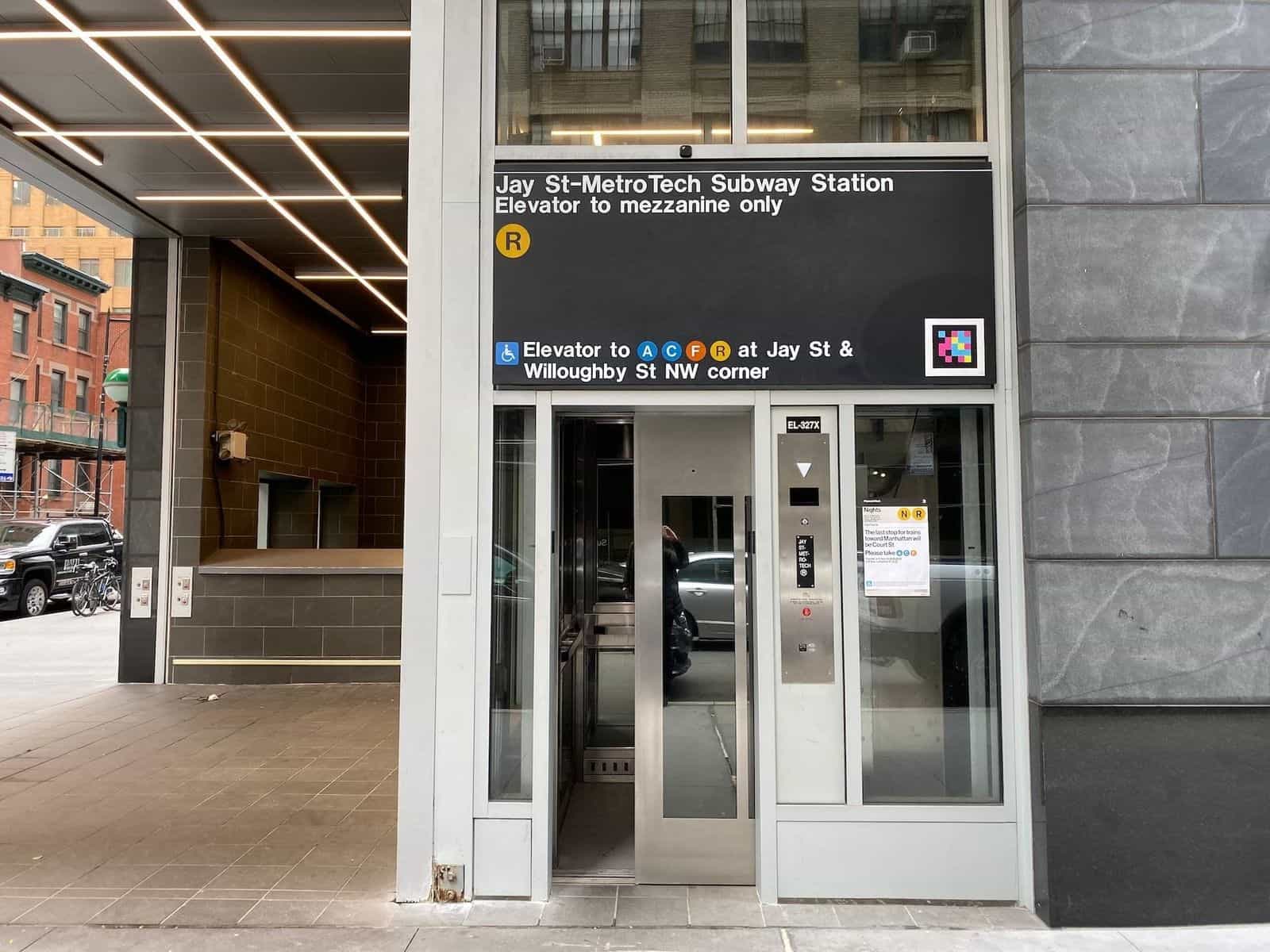 Outdoor elevator at Jay St-MetroTech on the northwest corner of Jay St & Willoughby St: stainless-steel glazed cabin and, above it, the sign «Jay St-MetroTech Subway Station · Elevator to mezzanine only» with the R bullet and a NaviLens code next to the message «Elevator to A C F R»