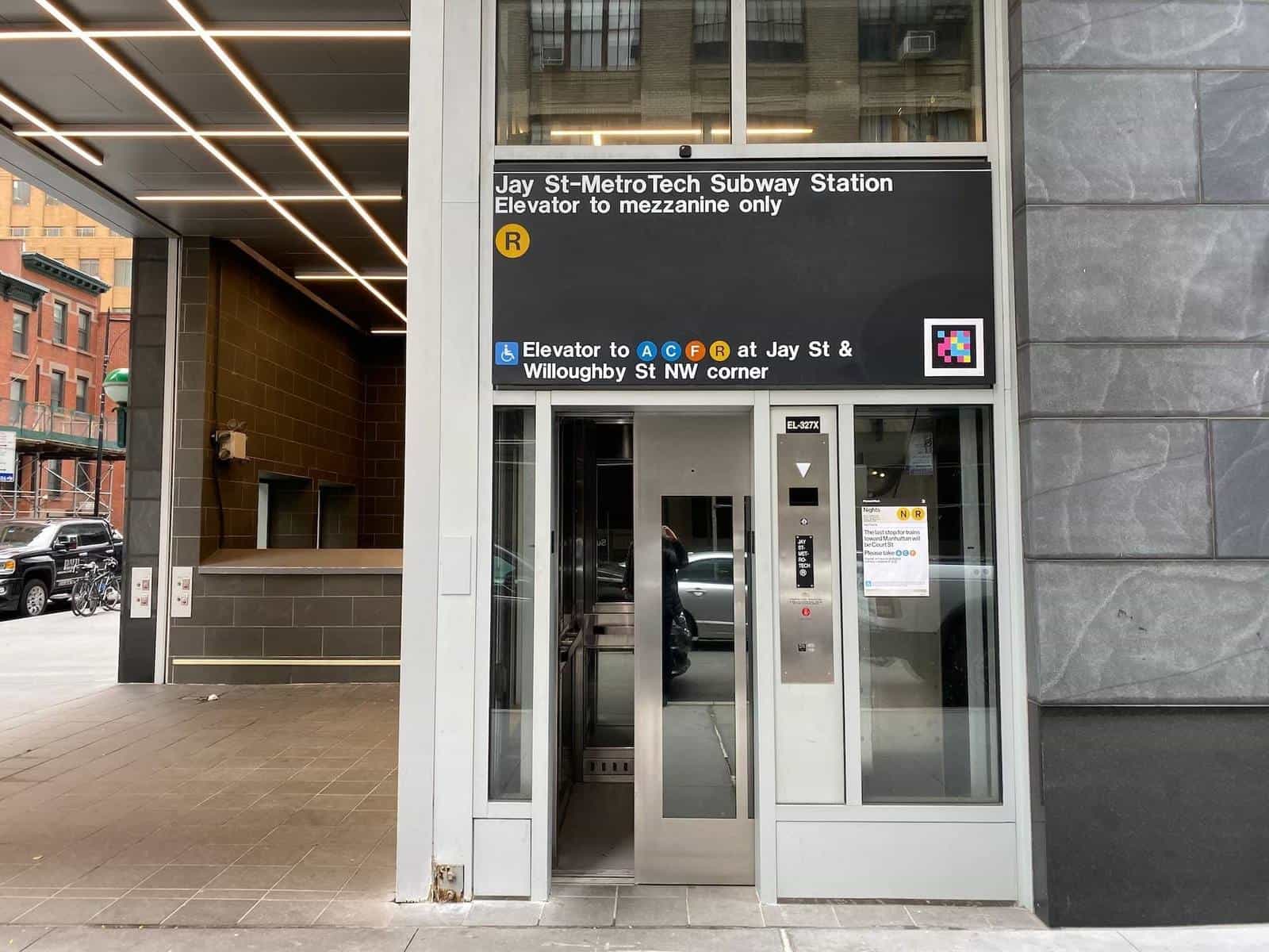 Outdoor lift at Jay St-MetroTech on the northwest corner of Jay St & Willoughby St: stainless-steel glazed cabin and, above it, the sign «Jay St-MetroTech Subway Station · Lift to mezzanine only» with the R bullet and a NaviLens code next to the message «Lift to A C F R»