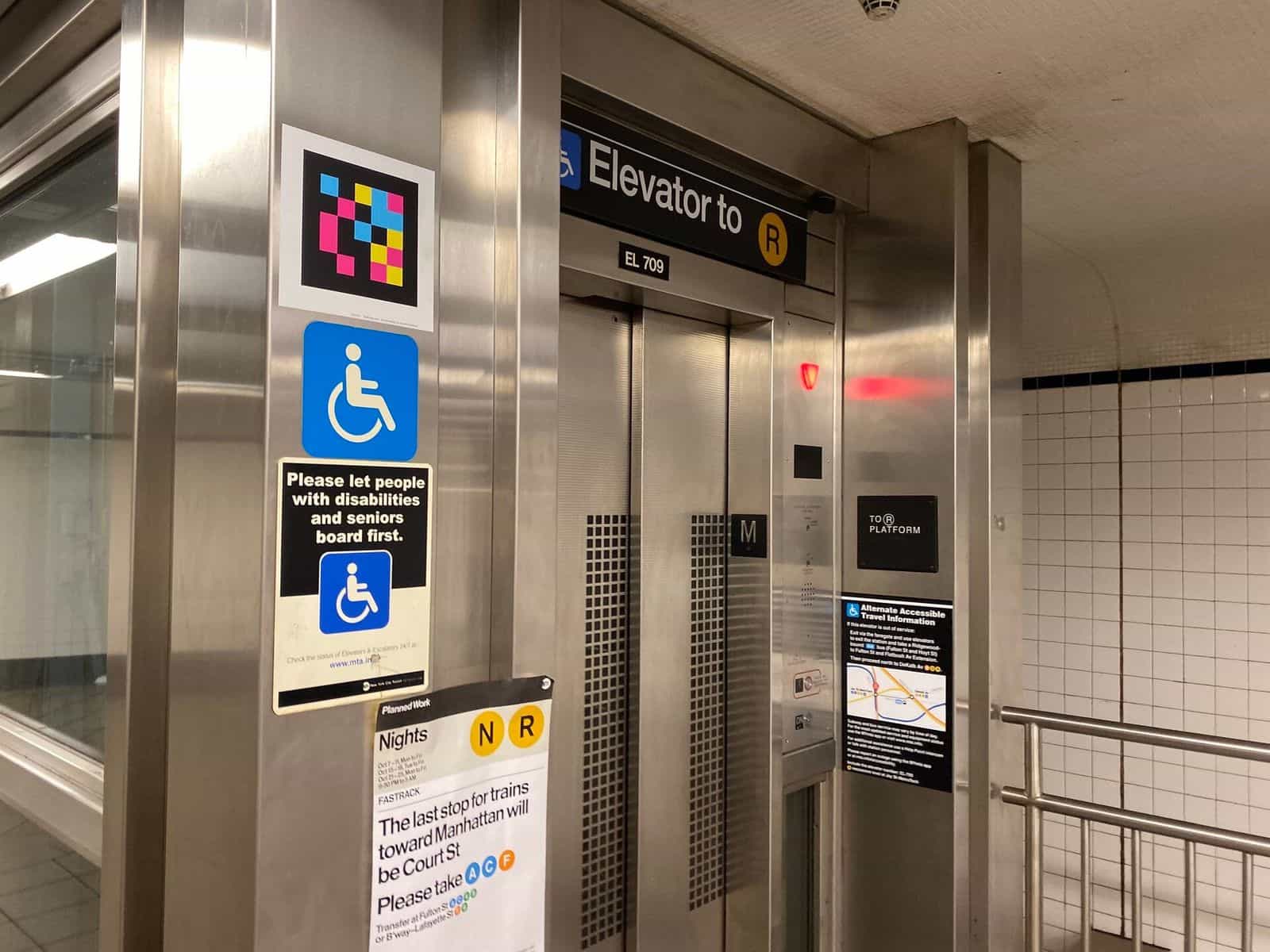 Elevator «EL 709 · Elevator to R» at Jay St-MetroTech: stainless-steel cabin with red warning light, blue wheelchair pictogram, sign «Please let people with disabilities and seniors board first» and a square NaviLens code at the top of the column; below, a «Planned Work — Nights N R» notice with rerouting via Court St / Fulton St / Lafayette St