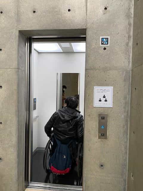 Person im Rollstuhl betritt den barrierefreien Aufzug des Vangi-Museums, mit einem NaviLens-Code an der Betonwand neben dem Ruftaster