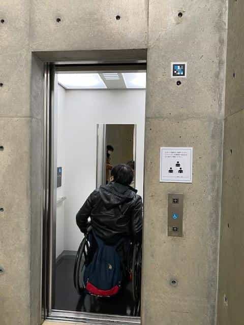 Person in a wheelchair entering the Vangi Museum's accessible lift, with a NaviLens code on the concrete wall next to the call button