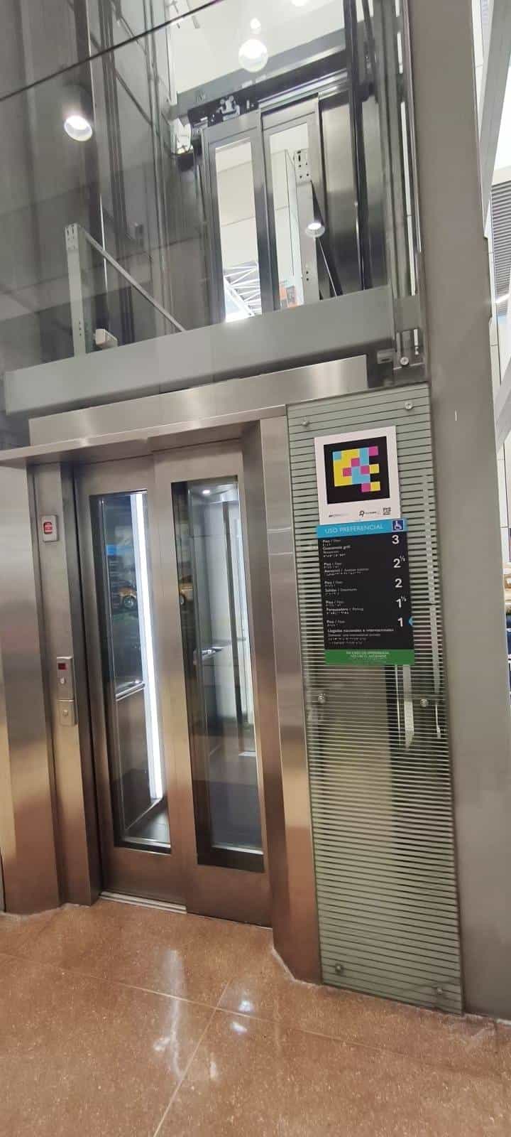 Glass-and-steel panoramic lift at José María Córdova Airport with a square NaviLens code in cyan, magenta and yellow above the blue “USO PREFERENCIAL” panel, which lists in Spanish, English, braille and Guarani the floors: 3 Guacamaya grill, 2½, 2 Aerocivil, 1½ Salidas/Departures, 1 Parqueaderos, 0 Domestic and international arrivals, with a green legend “In case of emergency, do not use the lift”