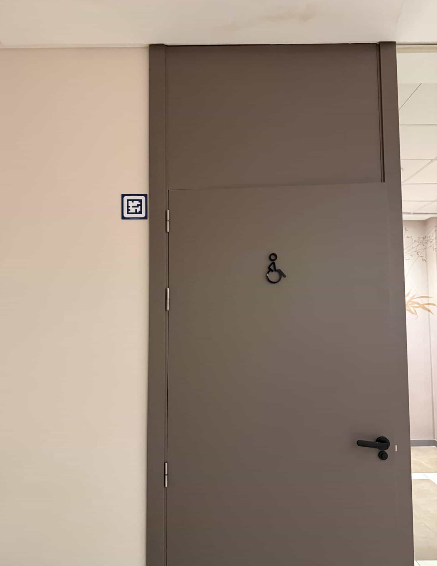 Warmgraue Tür des barrierefreien WC im AR Diamante Beach, mit schwarzem Rollstuhl-Piktogramm und blauem NaviLens-Code an der Wand