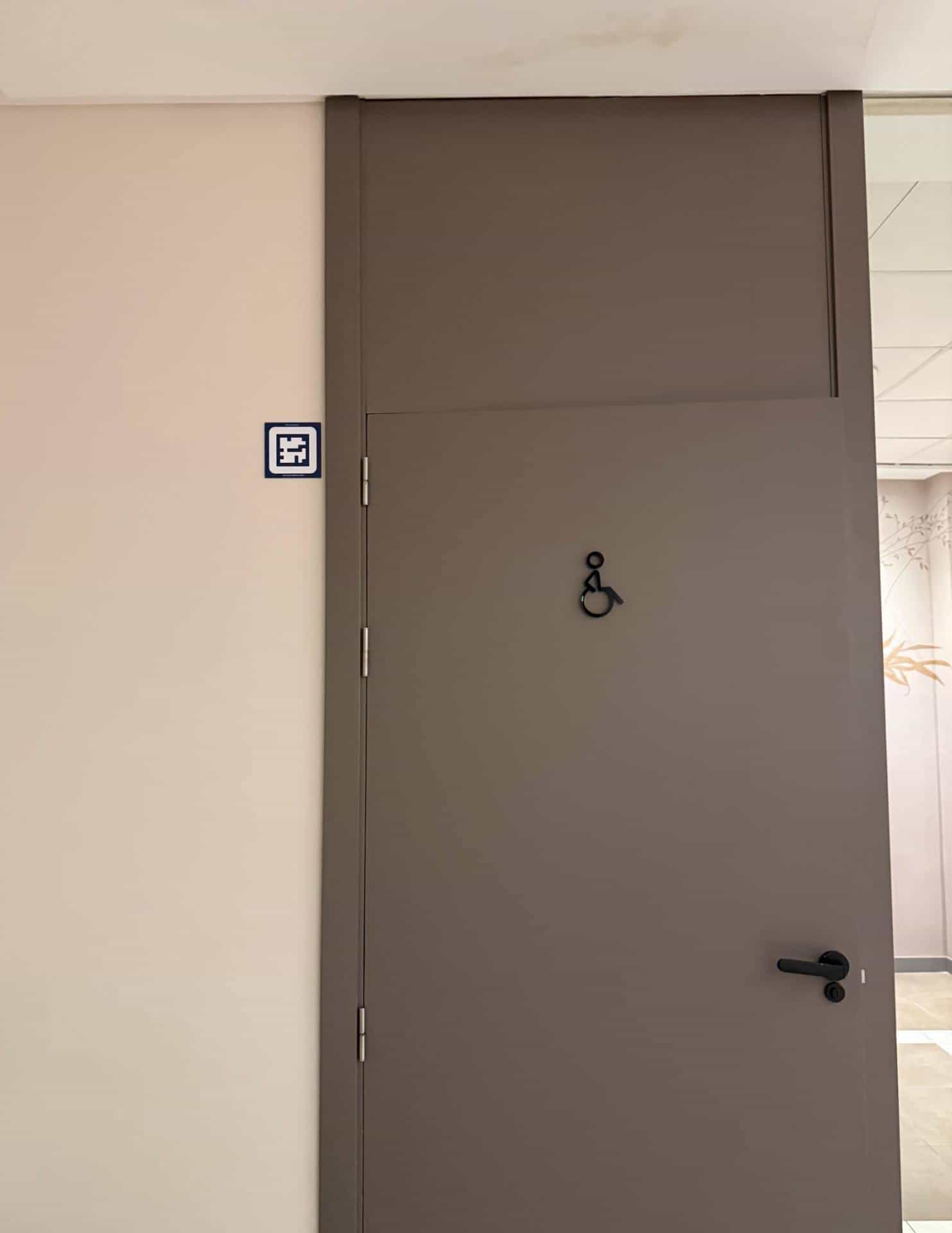Warm-grey door of the accessible toilet (wheelchair user) at the AR Diamante Beach, with a black wheelchair pictogram and a blue NaviLens code on the wall