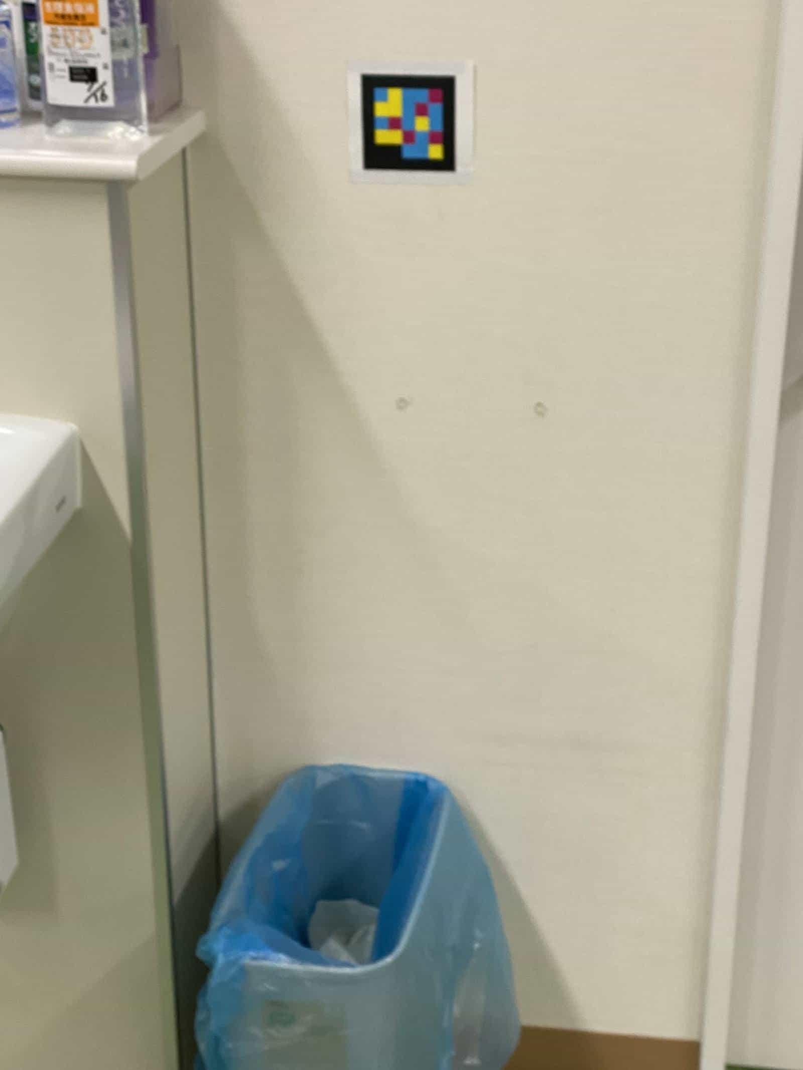 Detail of the department toilet: smaller laminated NaviLens code stuck on the cream wall, just above a bin with a translucent blue bag — size and height calibrated so a low-vision user can capture it on entering