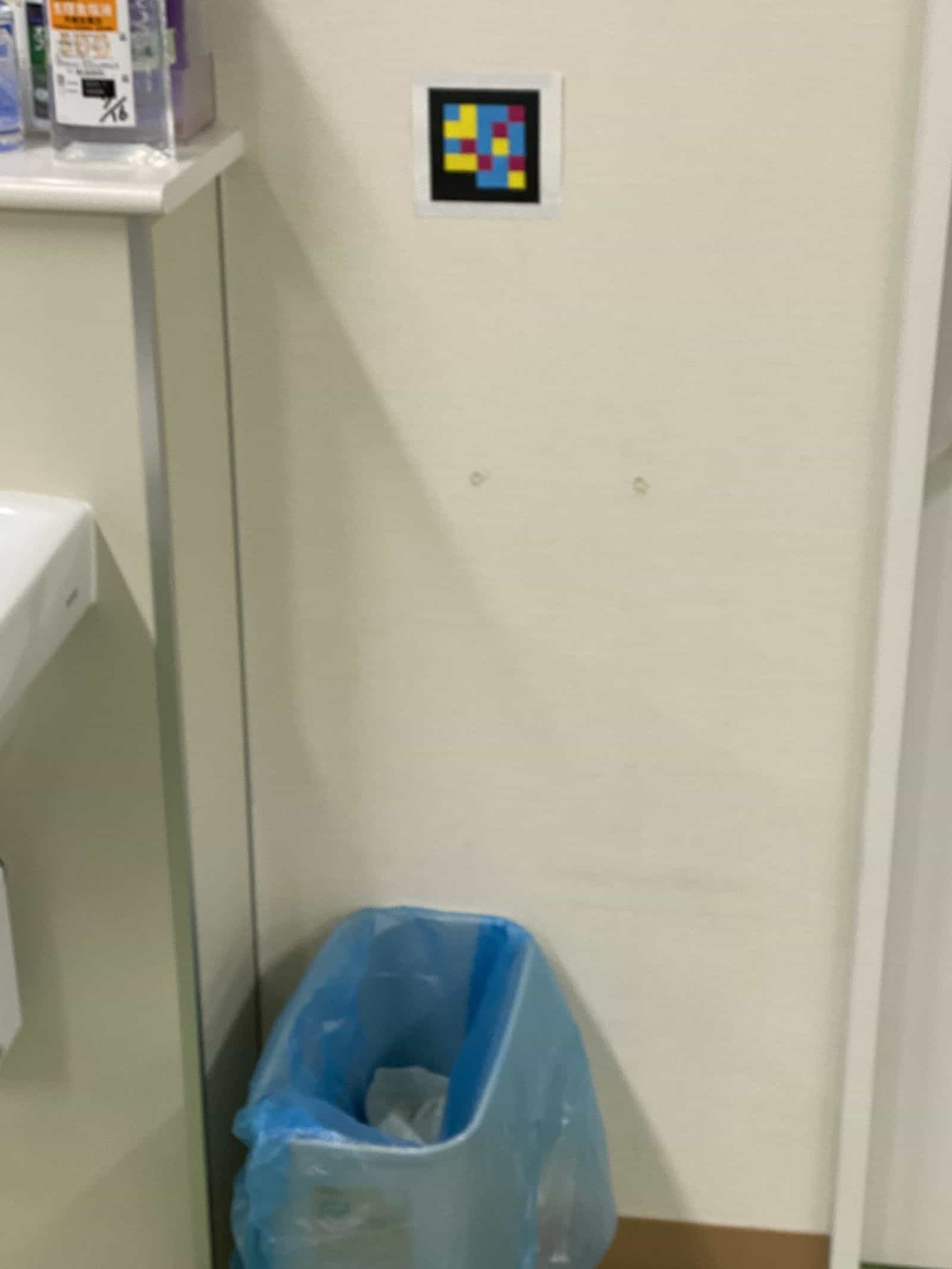 科のトイレの細部：クリーム色の壁に貼られた小さめのラミネート版NaviLensコード、半透明の青いごみ袋が入ったゴミ箱のすぐ上 — ロービジョン利用者が入室時に読み取れるよう、サイズと高さが調整されている
