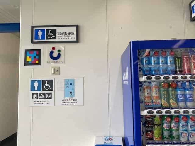 神戸空港駅の「男子お手洗 / Men's Toilets」入口にNaviLensタグ、「こうべ・だれでもトイレ」絵文字、トイレ触知図、右側に100円・500円飲料を扱う青いアサヒの自販機