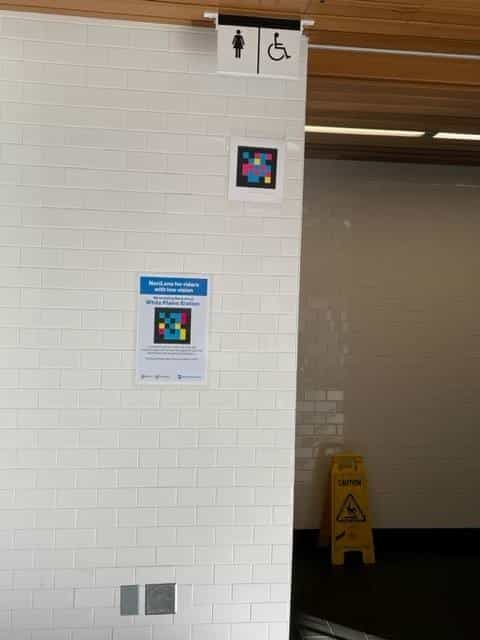 White tile wall next to the women's toilet at White Plains: above, the woman icon and the accessibility icon; in the middle, a NaviLens code framed in white; below, an official MTA blue sign reading “NaviLens for riders with low vision · We're testing NaviLens at White Plains Station”